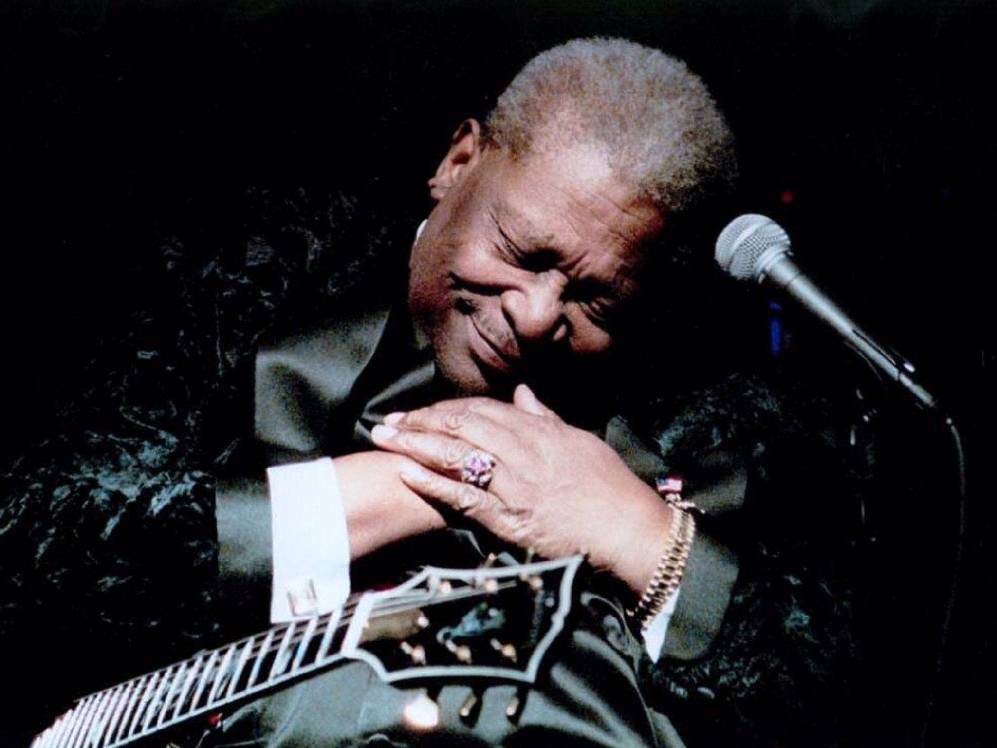 溫馨的吉他手B.B. King 壁紙 — 一位傳奇藍調音樂家在舞台燈光下熱情地彈奏結他，旁邊有一個咪高峰。