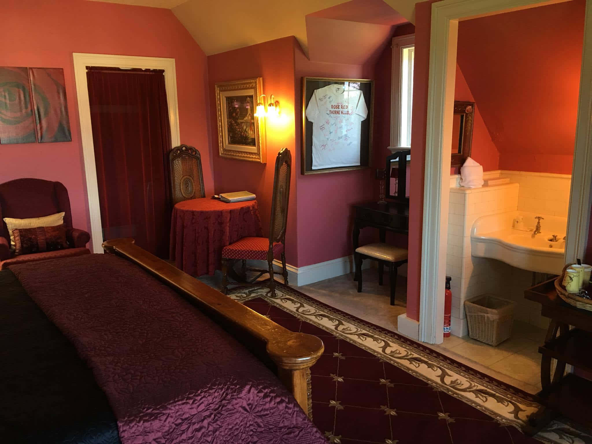 溫馨迷人的紅色房間 壁紙 — 一間燈光溫暖的復古臥室，擁有深紅色牆壁和豐富的紫色家具，配有木床、華麗地毯和相鄰的浴室。