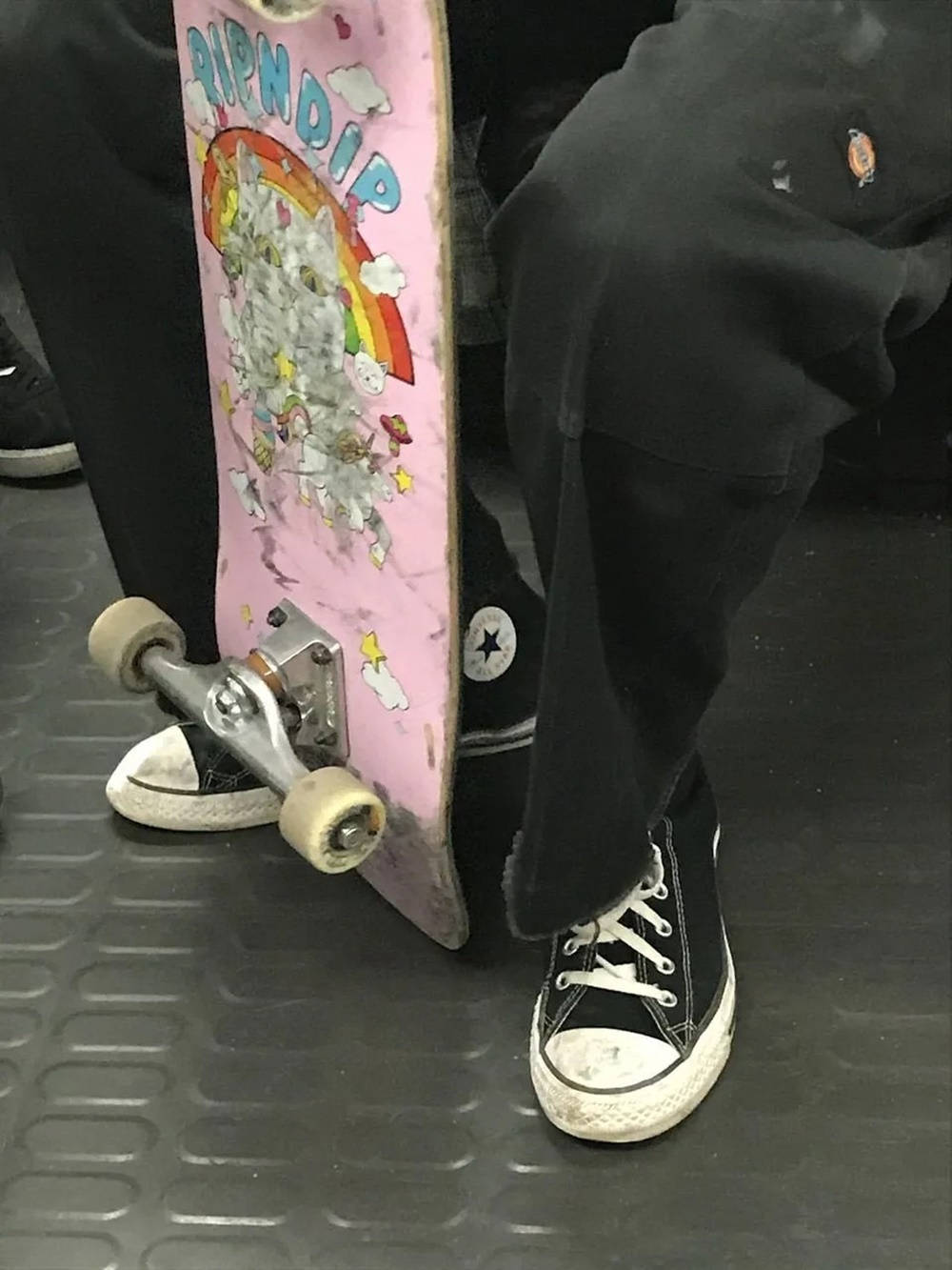 滑手搭乘火車 壁紙 — 一個人穿著黑色Converse運動鞋的腳部特寫，旁邊有一個粉紅色Ripndip滑板，輪子已磨損，放在有紋理的灰色地板上。
