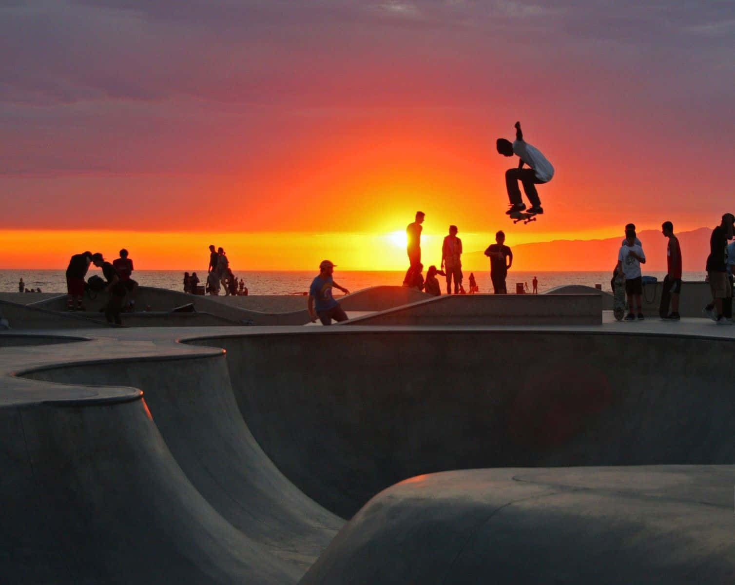 skateboarding 桌布 壁紙 — 滑板手在日落時分於海洋滑板公園上空做出空中技巧，背景是剪影人群。