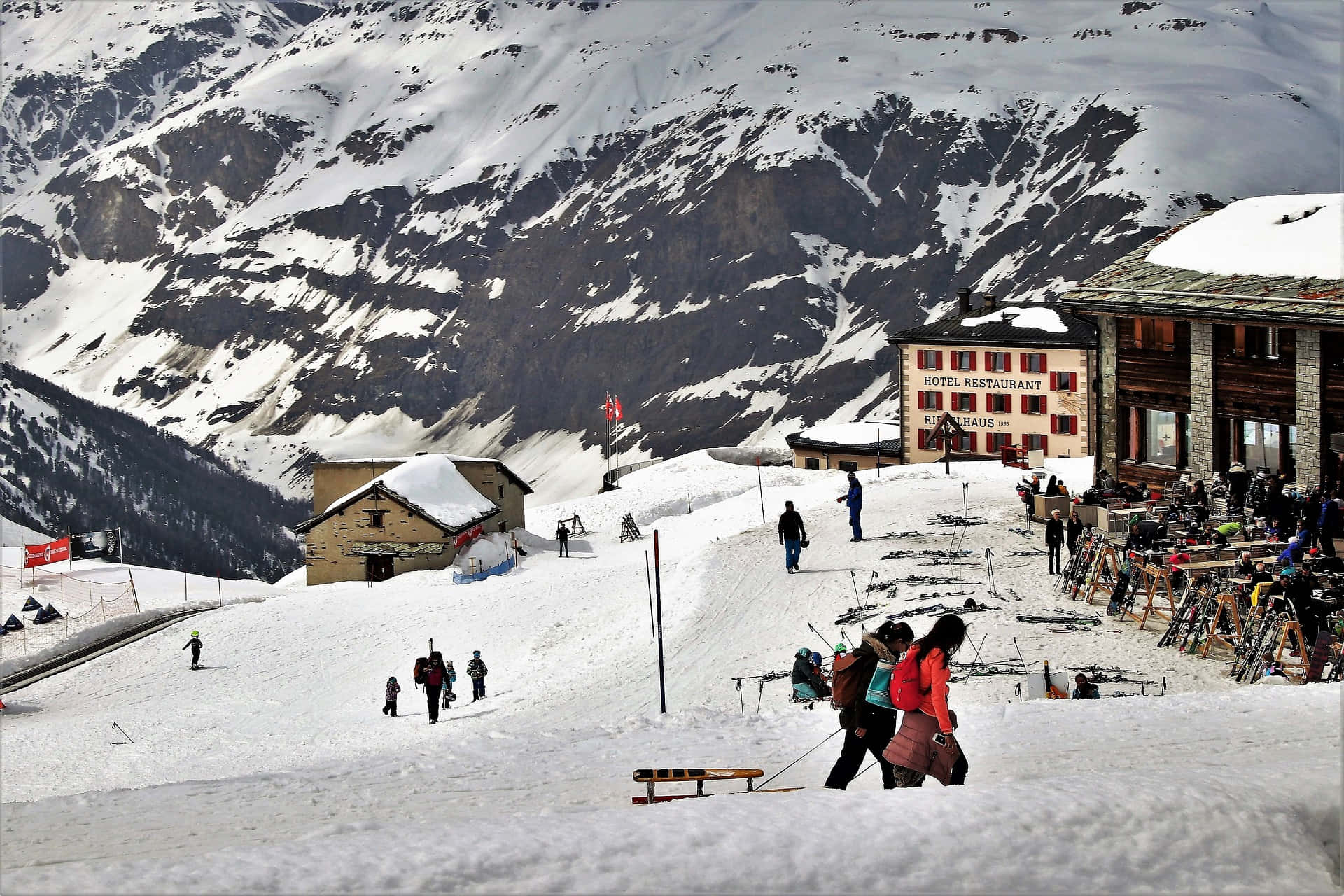 Ski Resort 桌布 壁紙 — 滑雪者和單板滑雪者聚集在山間小屋附近，背景是白雪覆蓋的山峰和岩石斜坡。