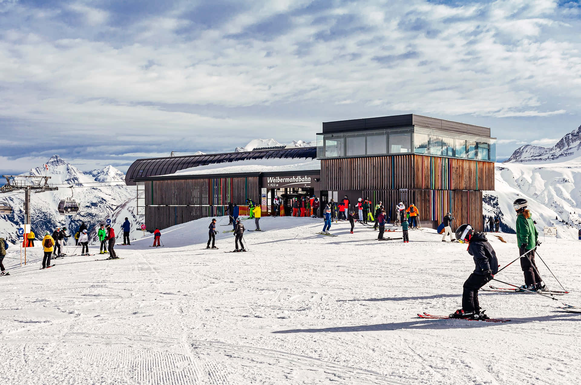 Ski Resort 桌布 壁紙 — 滑雪者和單板滑雪者聚集在現代化的山區車站，天空部分多雲，背景是白雪覆蓋的山峰。