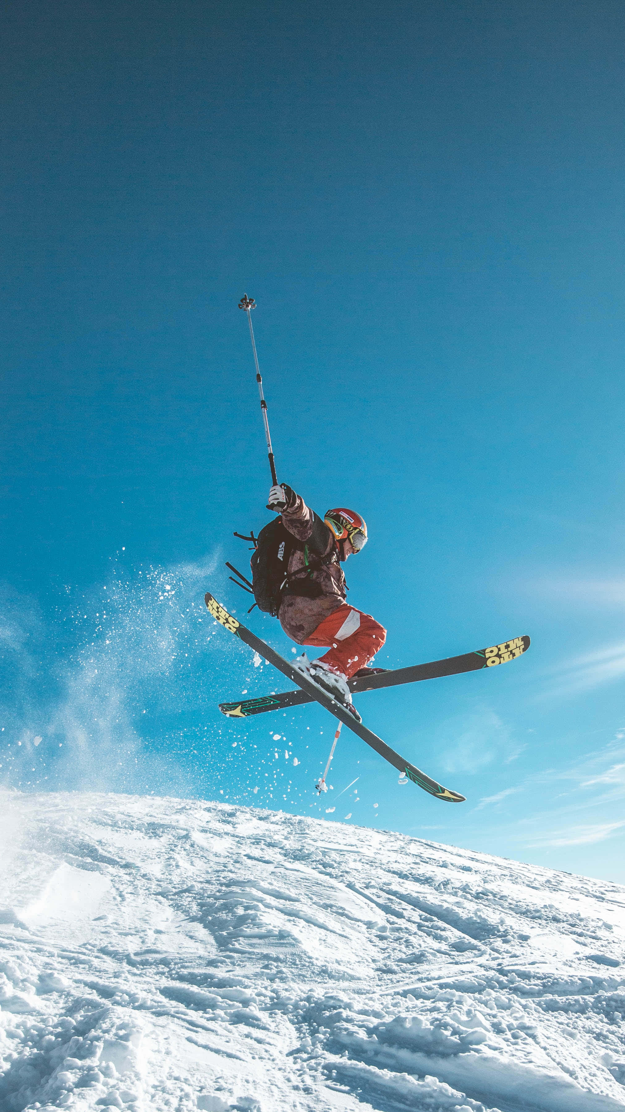 Skiing 桌布 壁紙 — 滑雪者在晴朗的藍天中飛翔，越過白雪覆蓋的斜坡，在明亮的陽光下揚起粉雪。