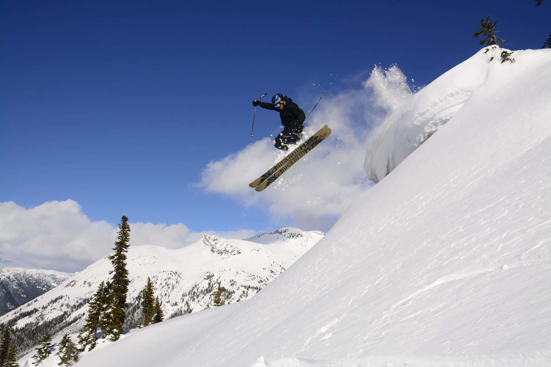 Skiing 桌布 壁紙 — 滑雪者在清澈的空氣中飛越白雪覆蓋的山峰，粉末狀的雪在明亮的藍天下飛濺。