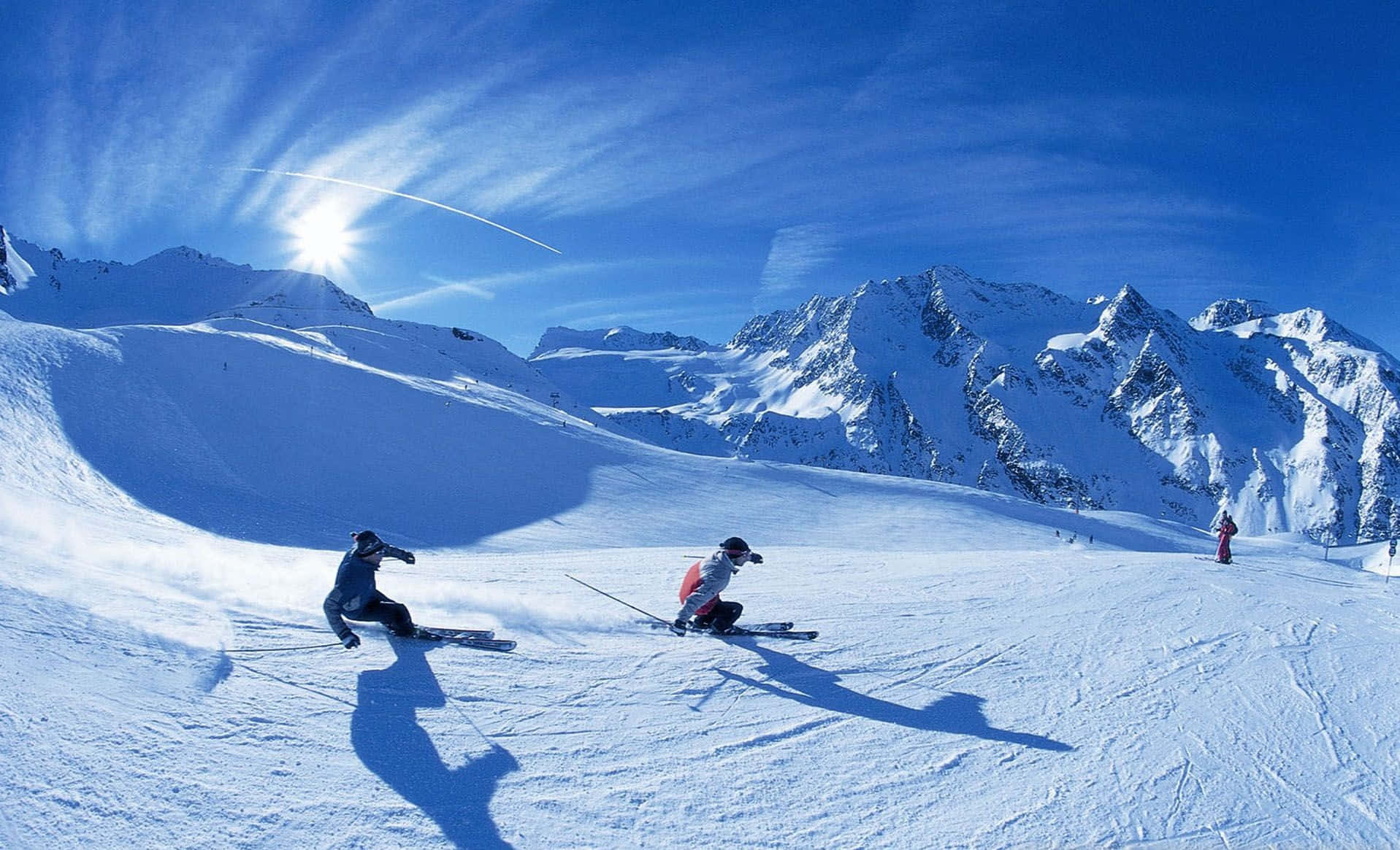 Winter Sports 桌布 壁紙 — 滑雪者在燦爛的陽光和山峰背景下，於鮮豔的藍天下劃過明亮的積雪。