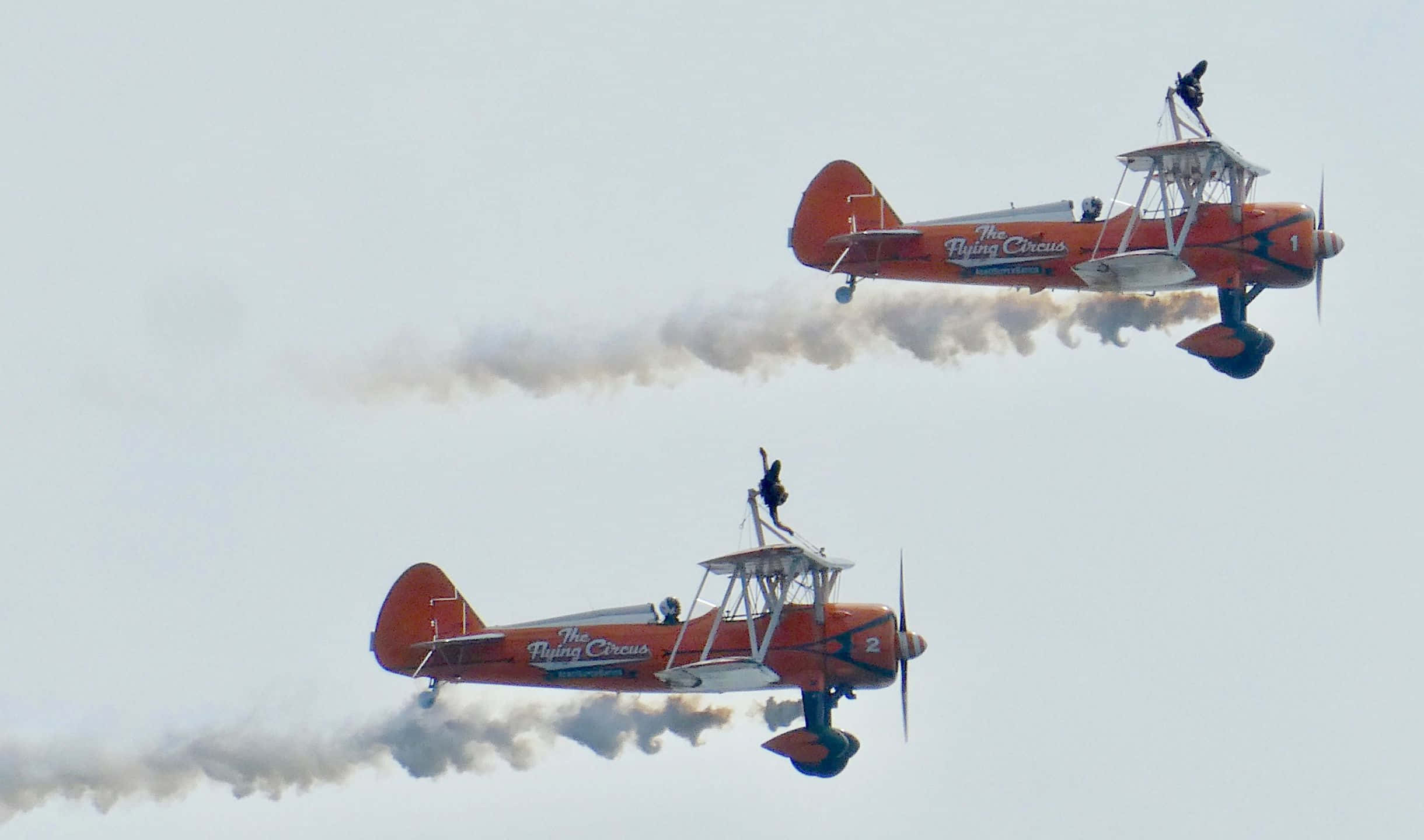 滿天歡樂 壁紙 — 兩架帶有「The Flying Circus」品牌的橙色雙翼飛機編隊飛行，在蒼白的天空中釋放煙跡。