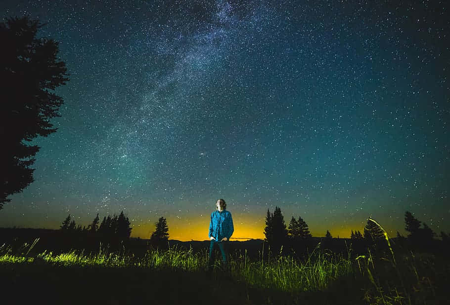 滿天繁星的夜空 壁紙 — 一個孤獨的人穿著藍色外套，站在草地上，頭頂是令人屏息的星空，銀河在閃耀。