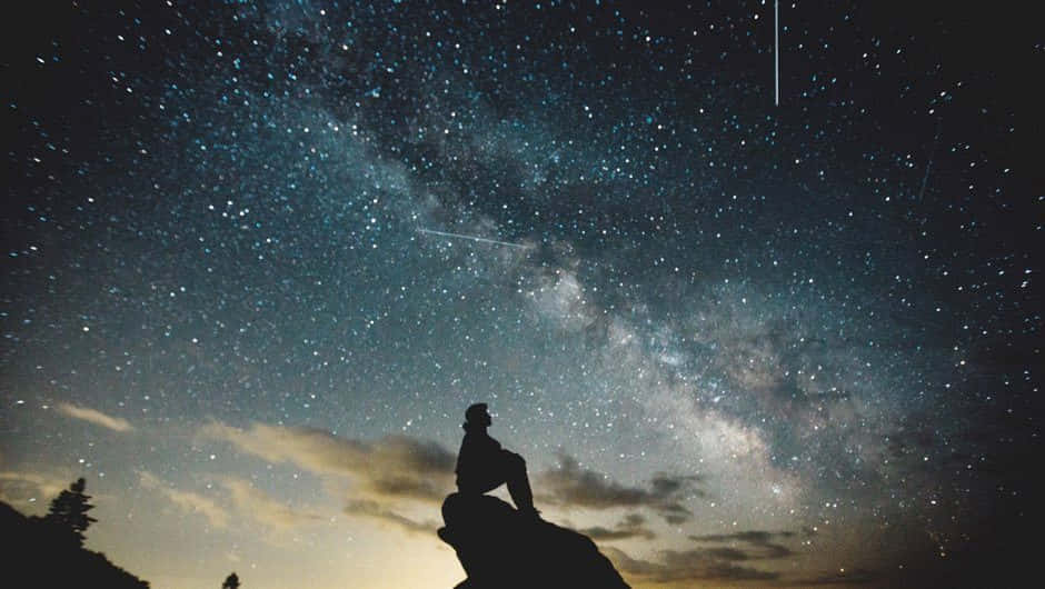 滿天閃爍的星星 壁紙 — 一個孤獨的身影坐在岩石露頭上，仰望充滿銀河和流星的壯麗星空。