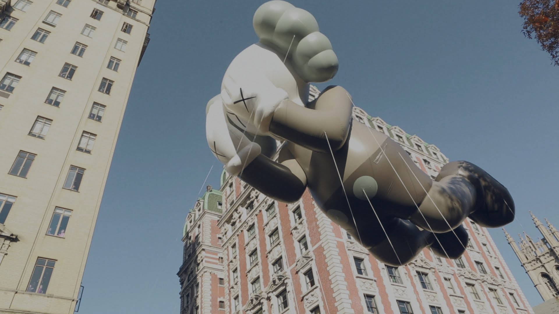 漂浮的KAWS Passing Through 壁紙 — 一個巨大的充氣KAWS熊在晴朗的藍天下漂浮在華麗的城市建築上方。