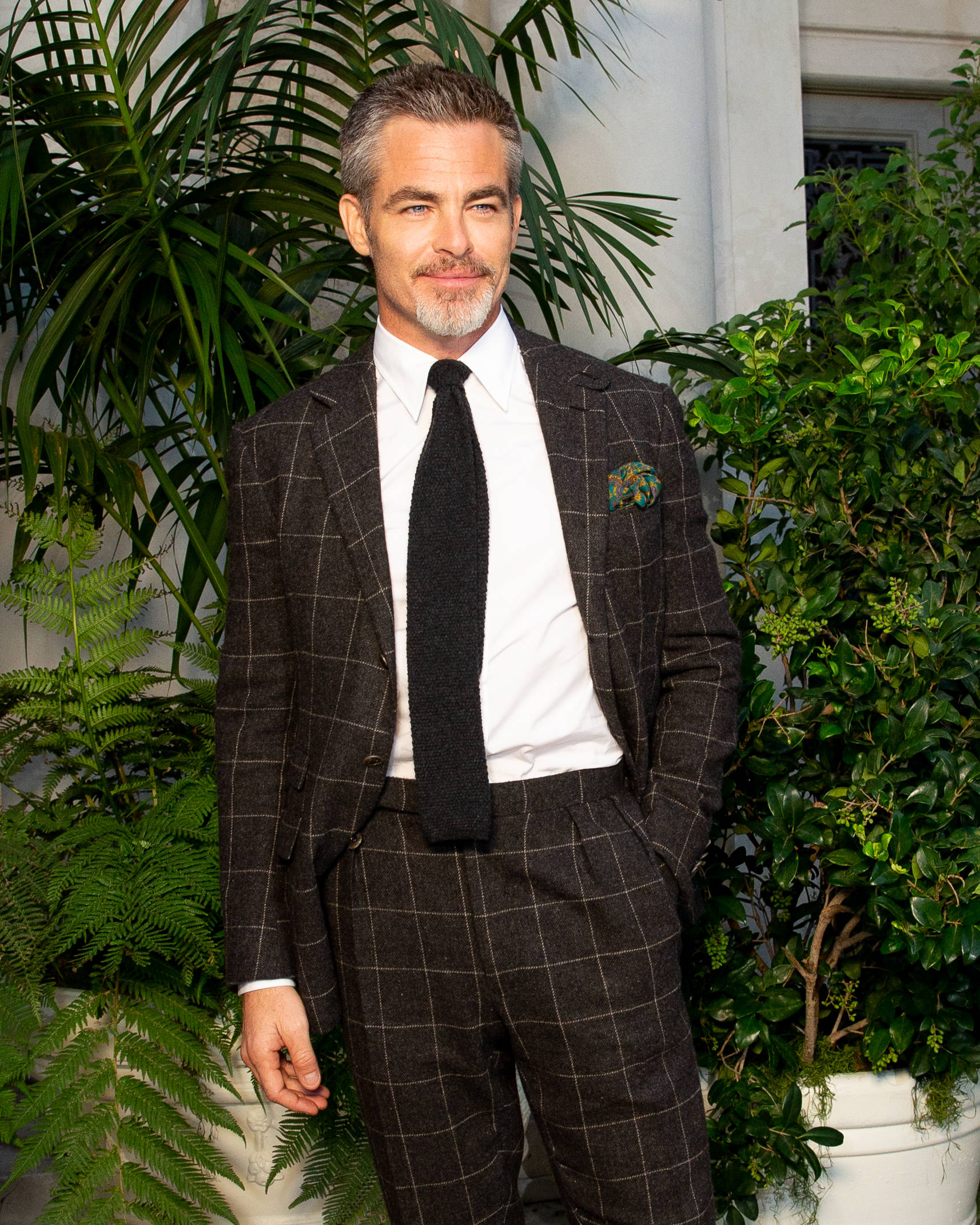 演員 Chris Pine 身著 Ralph Lauren 經典風範 壁紙 — 一位穿著格子西裝的時尚男士自信地站在茂盛的綠色盆栽植物中，背景是白色建築外牆。