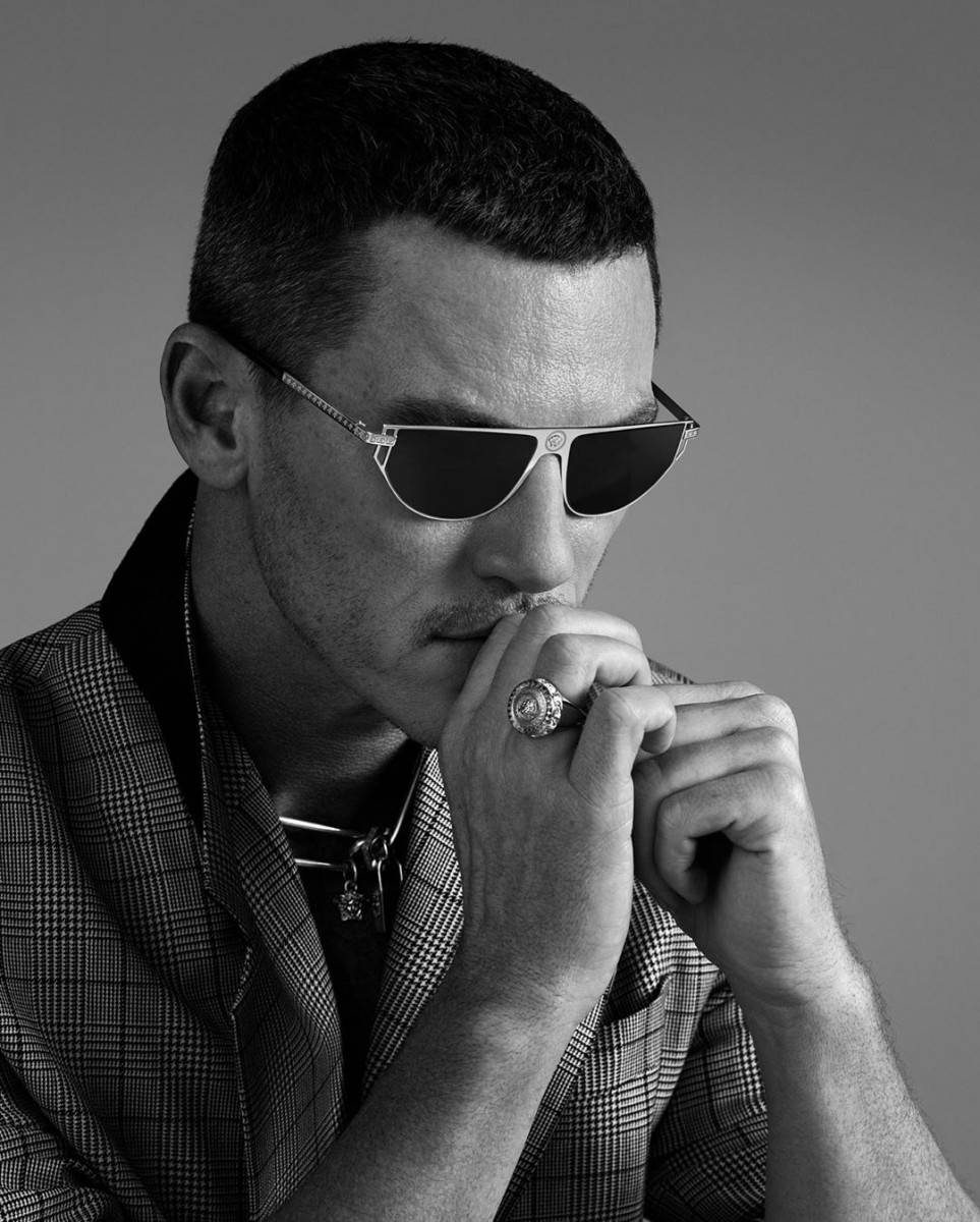 演員 Luke Evans 戴 Versace 太陽眼鏡 壁紙 — 一位戴著太陽眼鏡和格紋西裝外套的時尚男士，手指上戴著戒指，在灰色中性背景前沉思地擺姿勢。