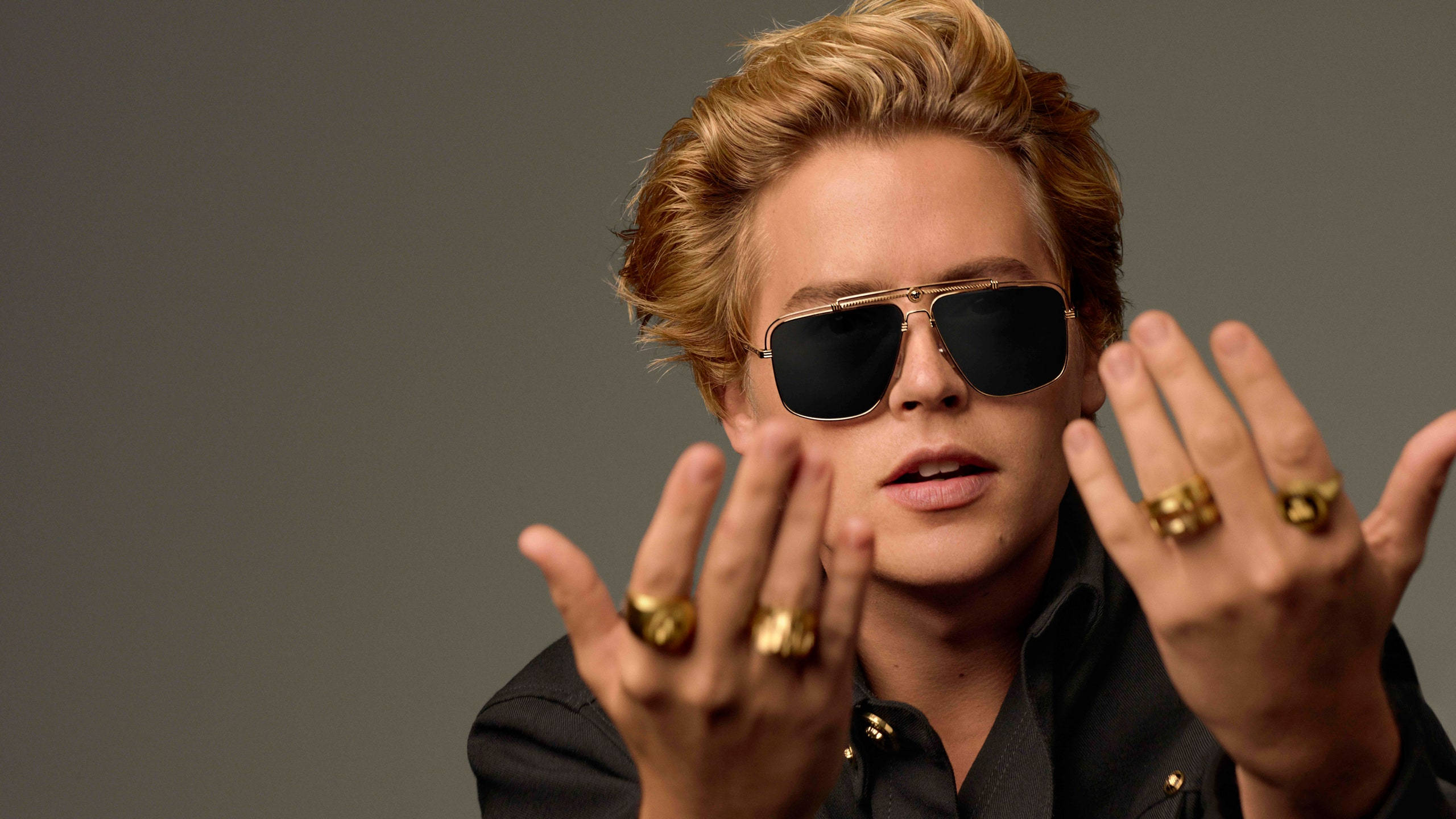 演員Cole Sprouse佩戴Versace太陽眼鏡 壁紙 — 一位時尚的金髮男子戴著飛行員太陽眼鏡，雙手戴著金戒指，在灰色中性背景前擺姿勢。