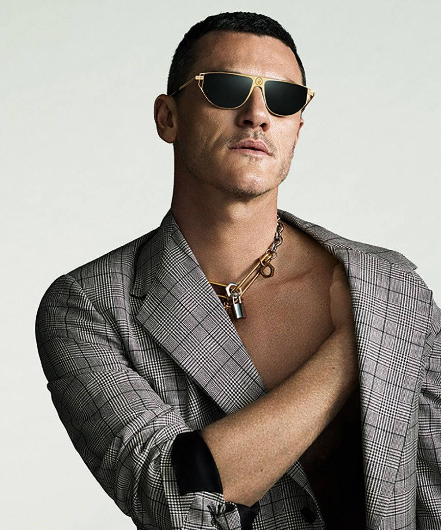 演員Luke Evans佩戴Versace眼鏡 壁紙 — 一位時尚男士穿著格紋西裝外套和金色太陽眼鏡，在純色背景前自信地擺姿勢。