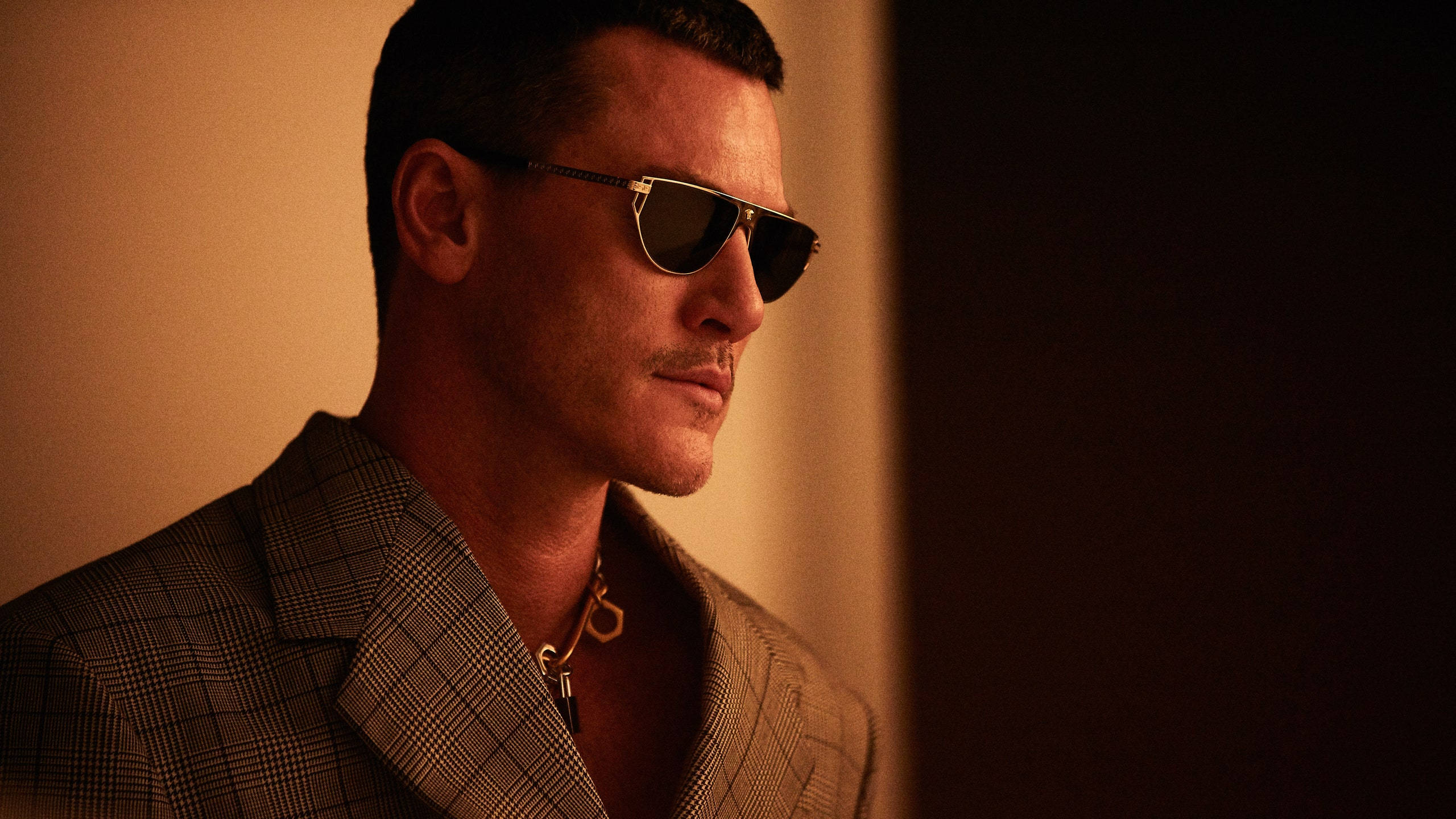 演員Luke Evans佩戴Versace太陽眼鏡系列 壁紙 — 一名身穿格紋西裝外套、戴著飛行員太陽眼鏡的男子，在溫暖的金色光線下站在深色背景前。