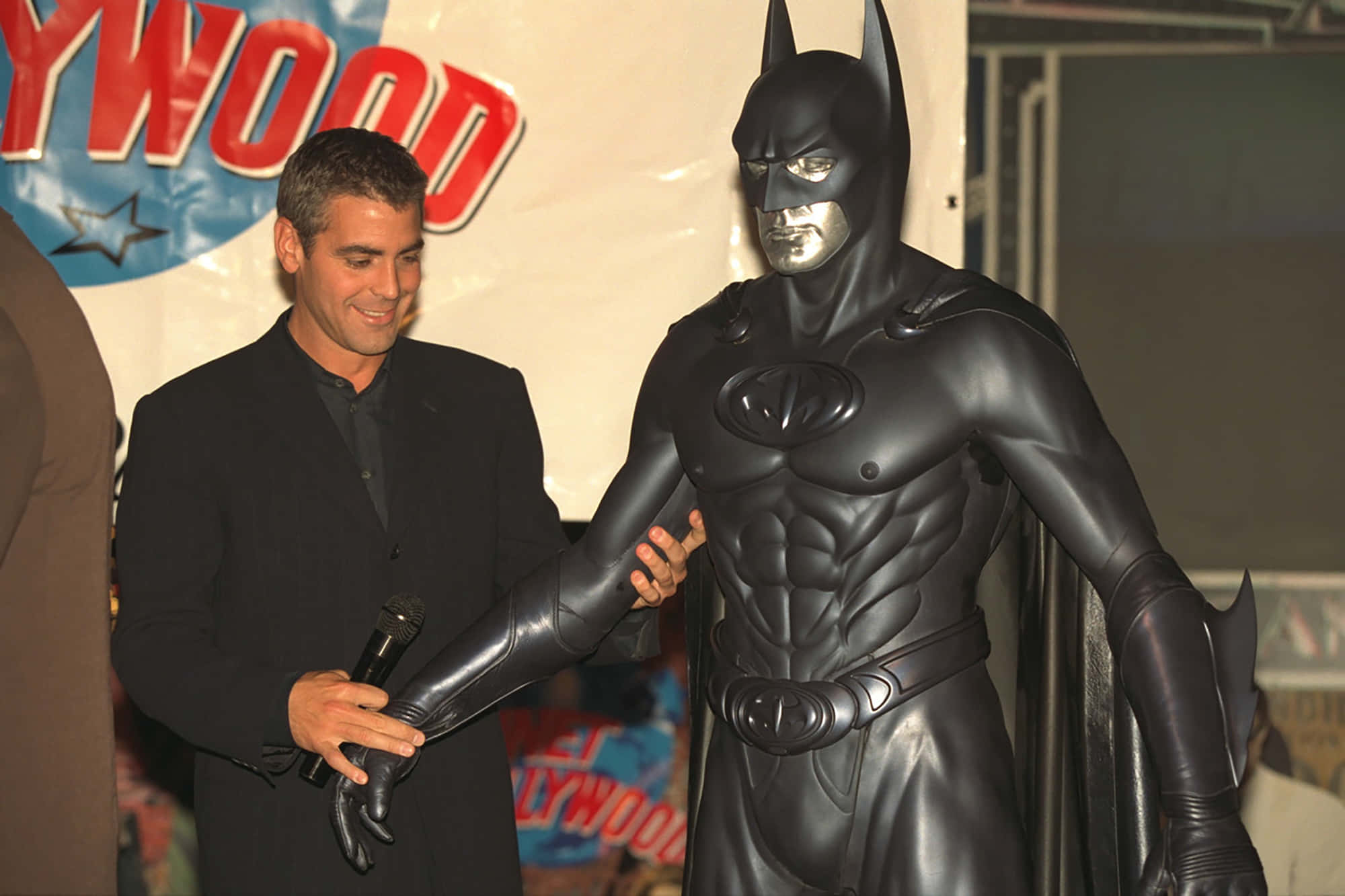 Bat-suit 桌布 壁紙 — 演員佐治古尼在荷里活活動中微笑著，手持麥克風站在一個真人大小的蝙蝠俠雕像旁。