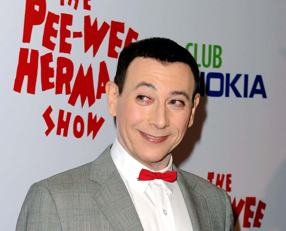 Paul Reubens 桌布 壁紙 — 演員兼喜劇演員 Paul Reubens 身穿灰色西裝和紅色領結，在「Pee-wee Herman Show」活動中微笑。