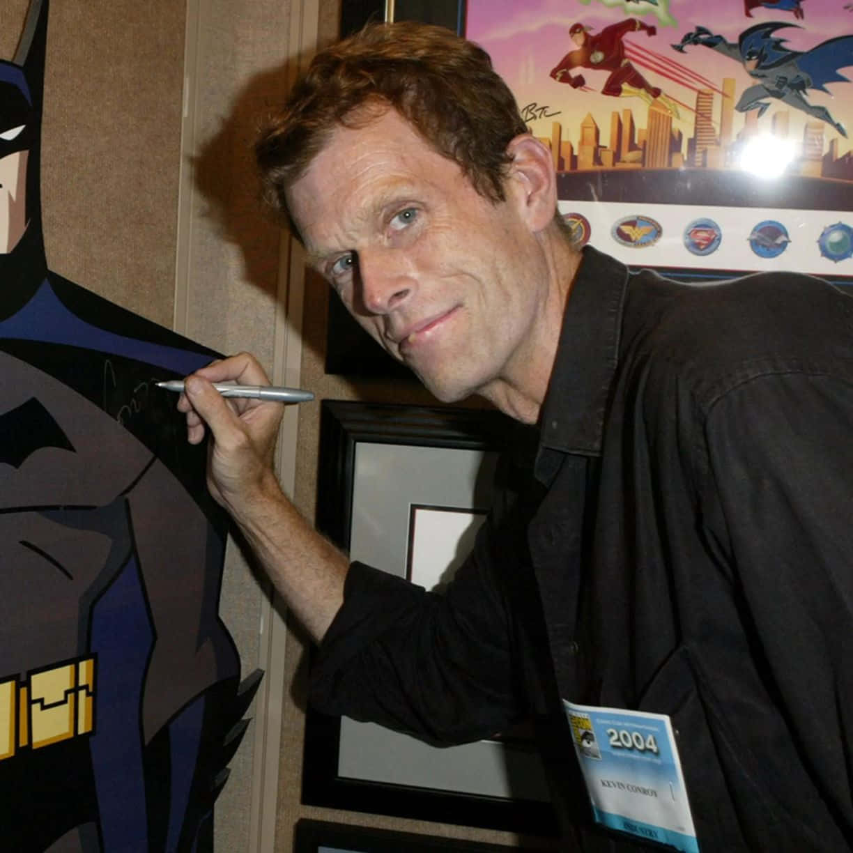 Kevin Conroy 桌布 壁紙 — 演員凱文·康羅伊在2004年漫畫展上，身後有超級英雄海報，為蝙蝠俠剪紙簽名。