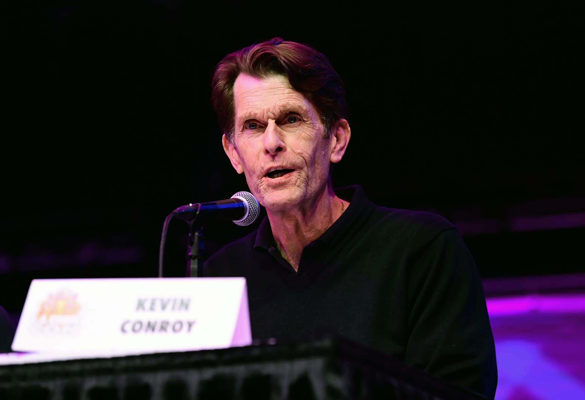Kevin Conroy 桌布 壁紙 — 演員凱文·康羅伊在一個燈光昏暗、帶有紫色舞台燈光的活動中對著麥克風講話。