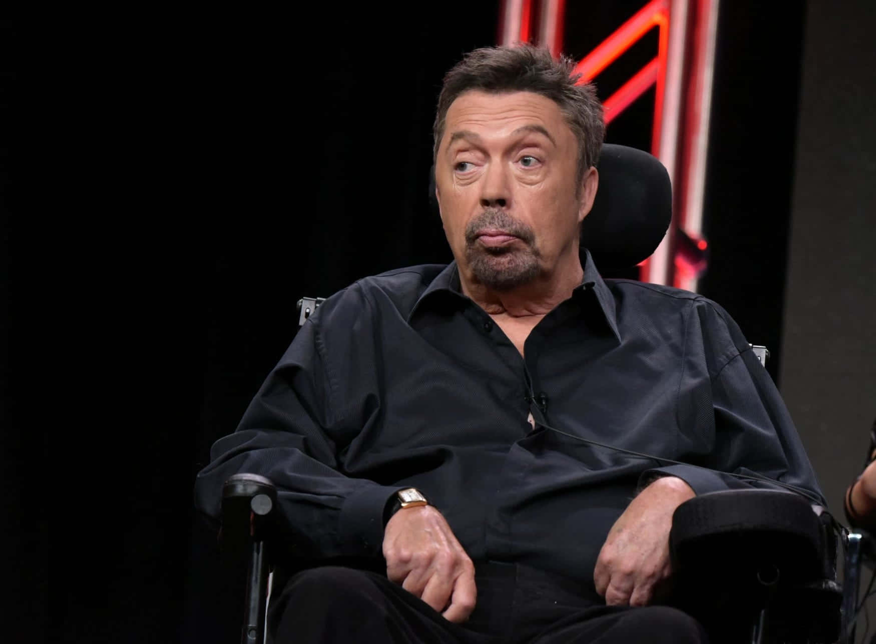 Tim Curry 桌布 壁紙 — 演員加利奧文坐在黑色椅子上，背景昏暗，身後有紅色霓虹燈光。