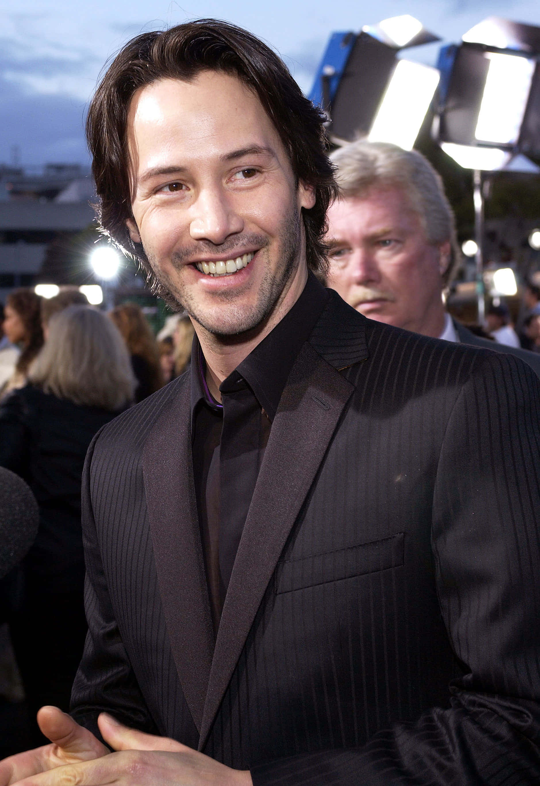 Keanu Reeves 桌布 壁紙 — 演員奇洛·李維斯在明亮的燈光下，穿著深色細條紋西裝，在戶外活動中溫暖地微笑。