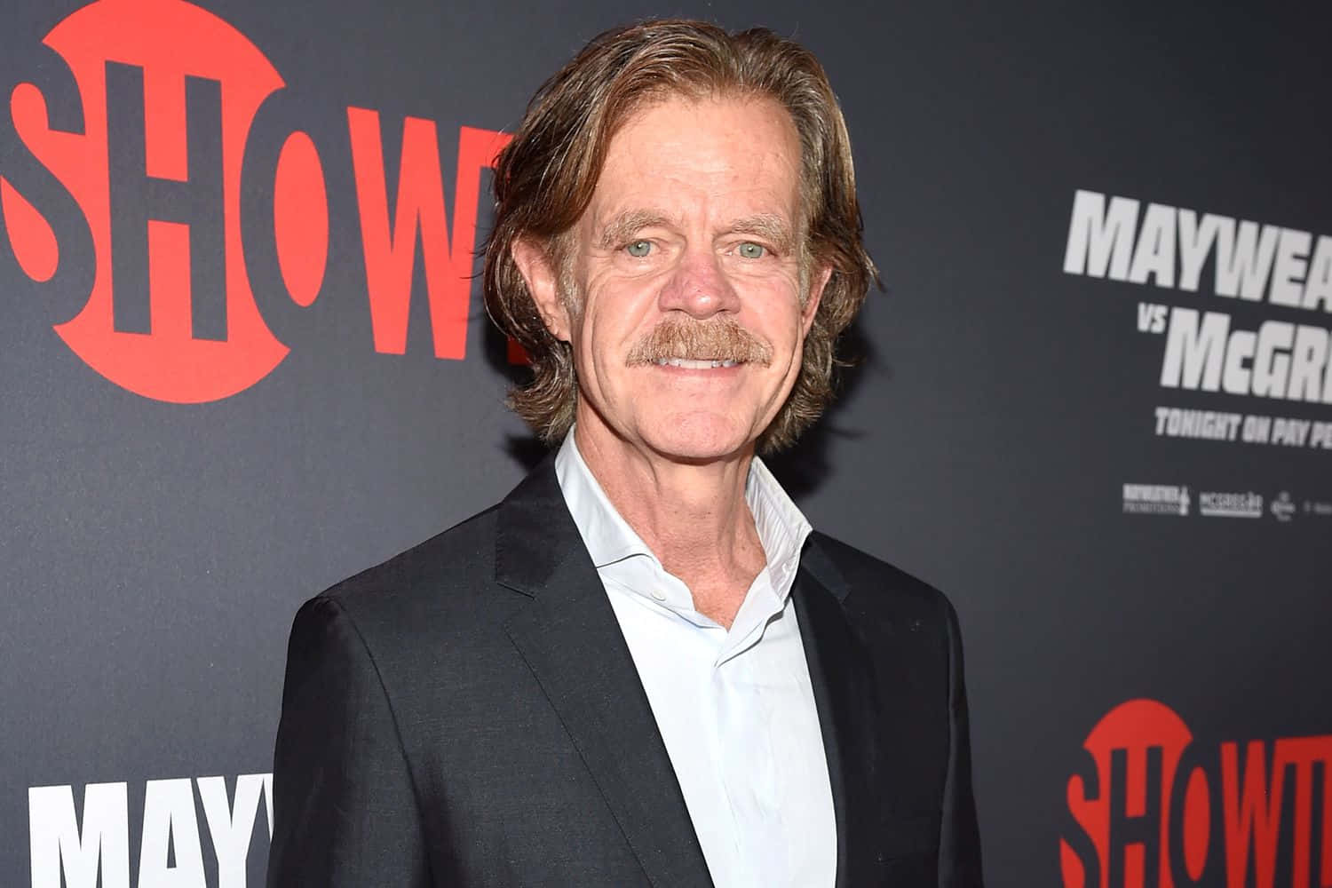 william h. macy 桌布 壁紙 — 演員威廉·H·梅西穿著黑色西裝和白色襯衫，在帶有紅色和白色活動品牌的深色背景前微笑著看向鏡頭。