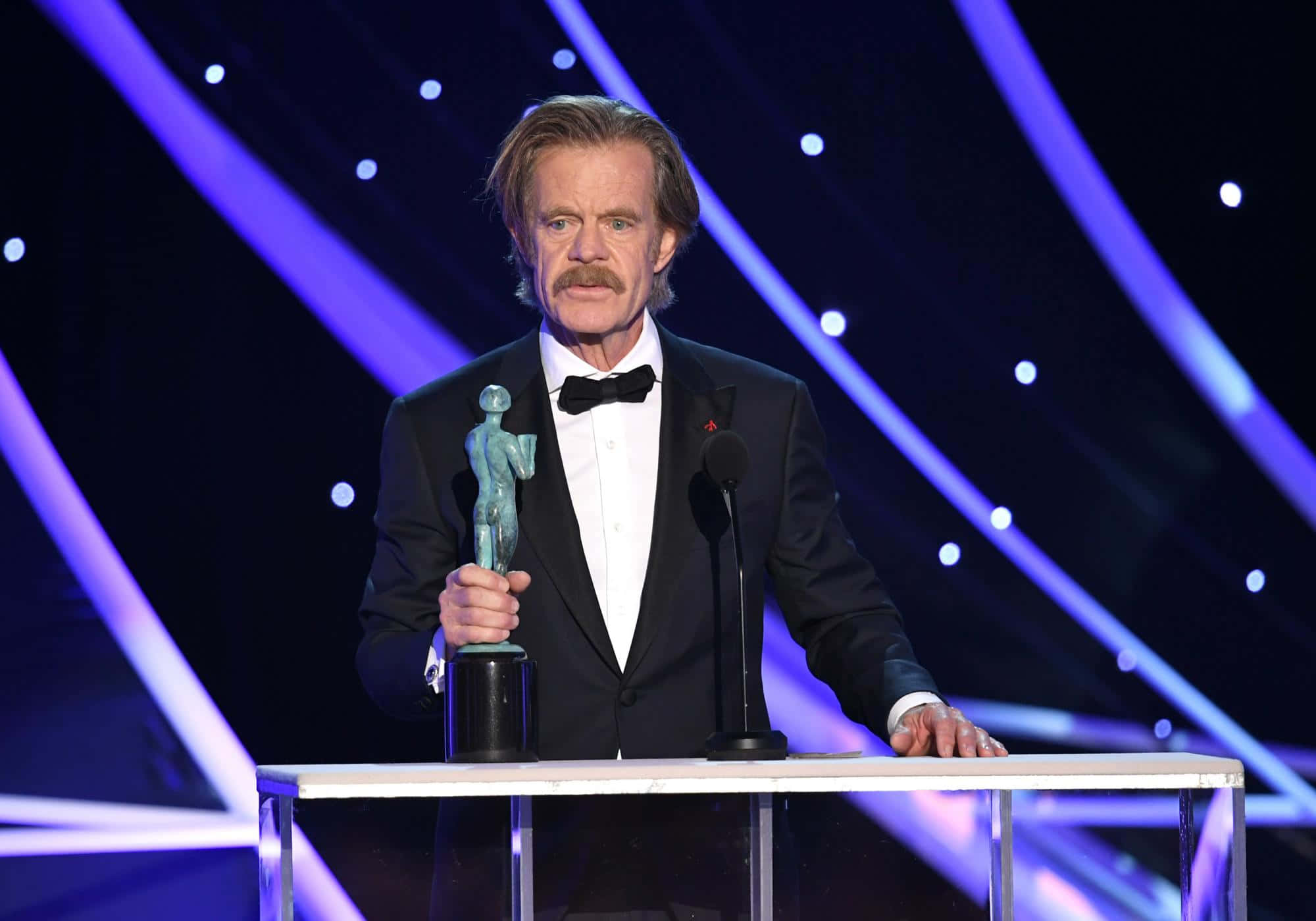 william h. macy 桌布 壁紙 — 演員威廉·H·梅西站在講台上，在戲劇性的紫色舞台燈光下手持獎項。