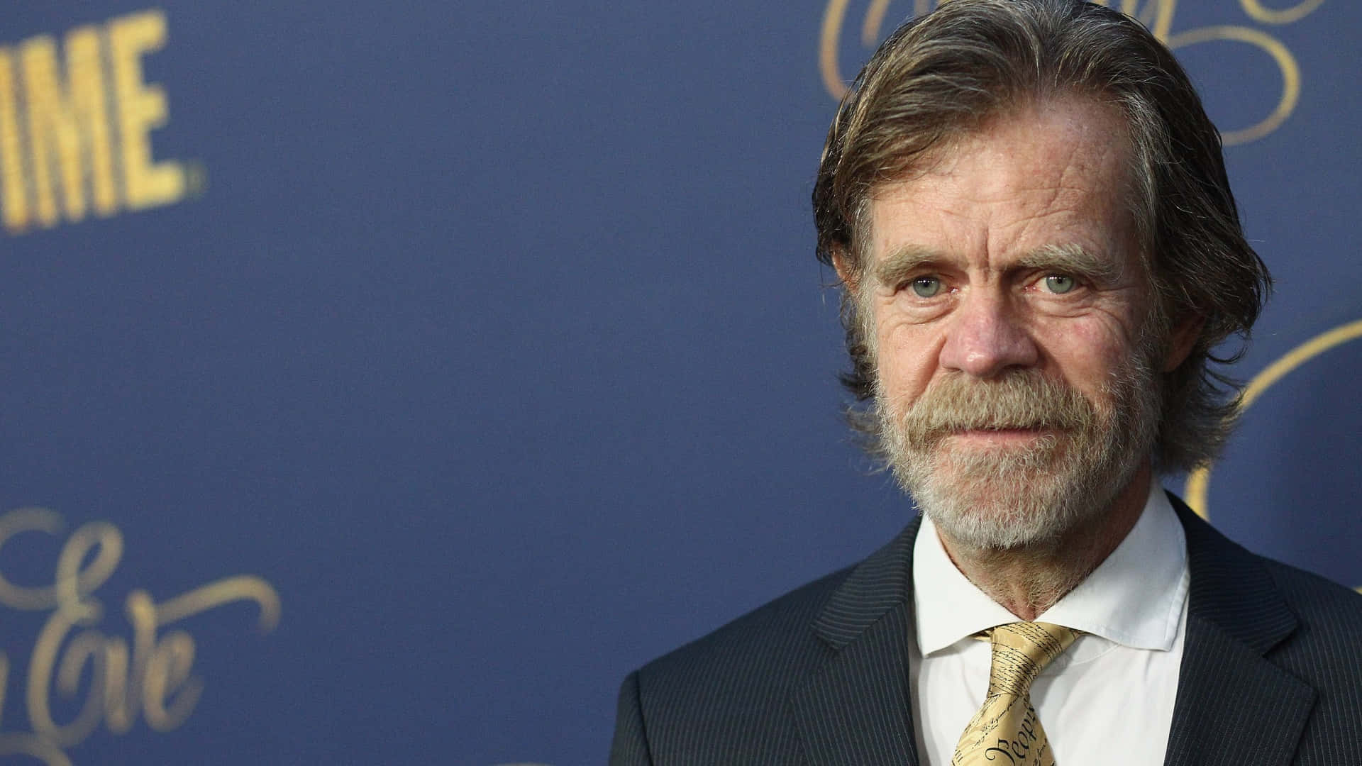 william h. macy 桌布 壁紙 — 演員威廉·H·梅西身穿深色西裝和金色領帶，在帶有金色字樣的藍色活動背景前擺姿勢。