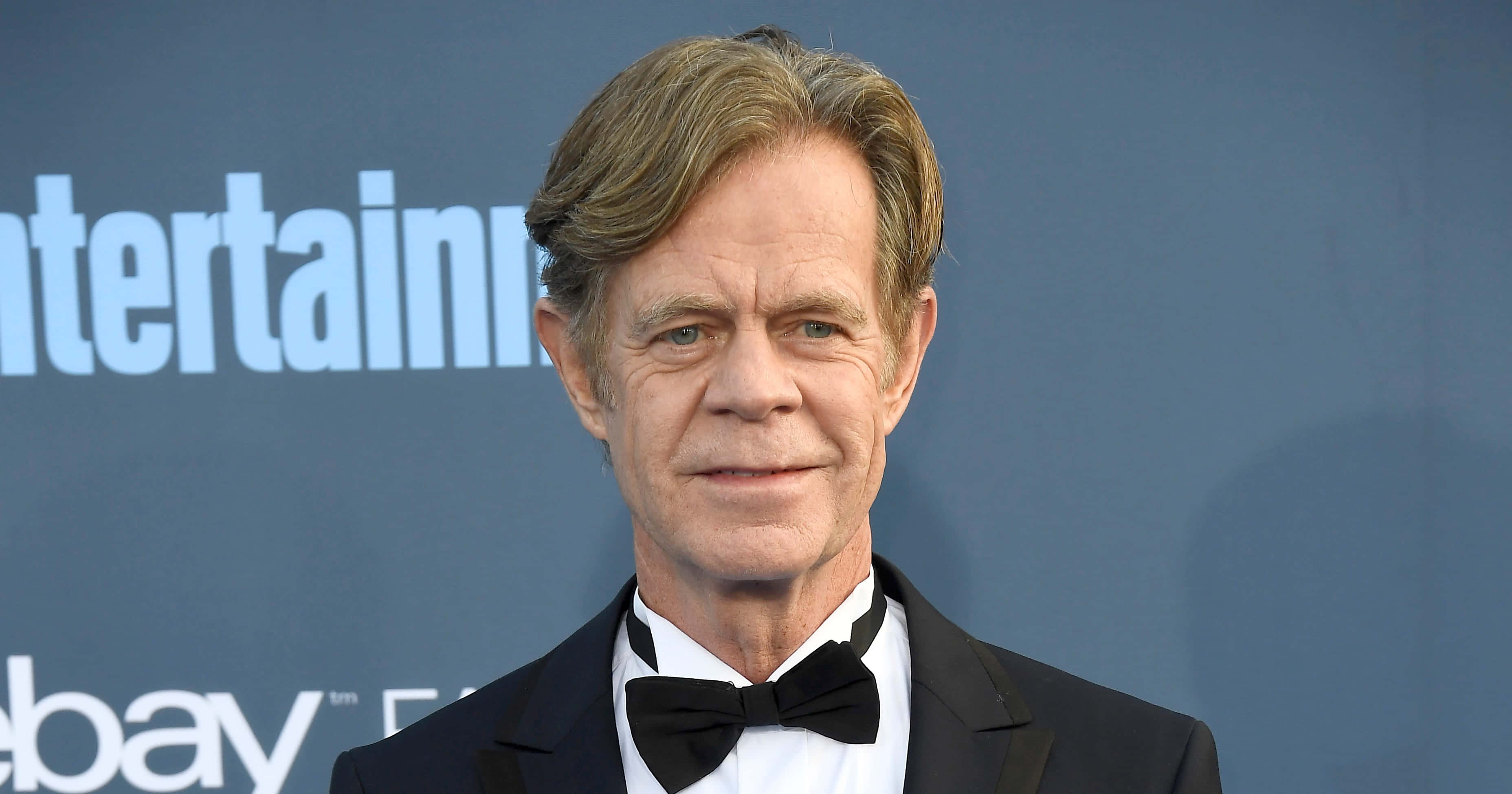 william h. macy 桌布 壁紙 — 演員威廉·H·梅西身穿黑色燕尾服和領結，在藍色品牌背景前自信地擺姿勢，出席娛樂活動。