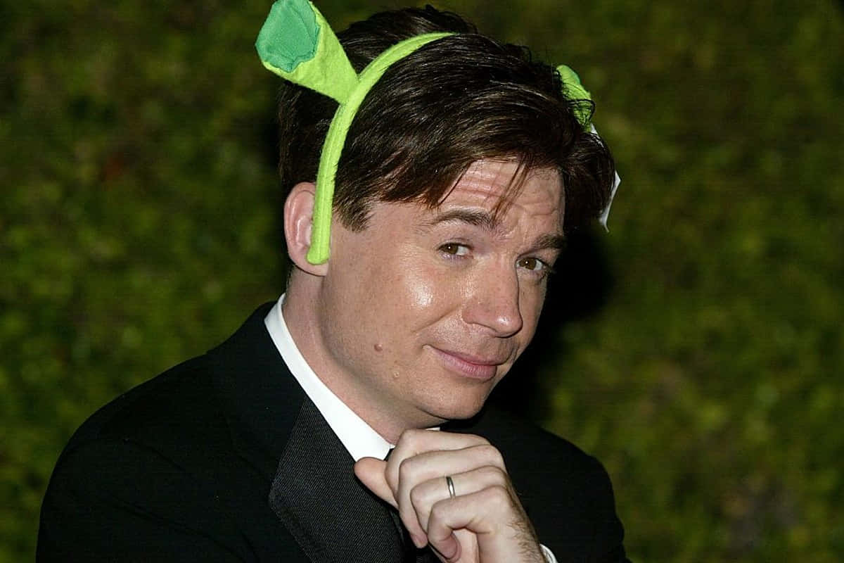 Mike Myers 桌布 壁紙 — 演員戴著綠色植物主題頭帶，穿著正式黑色西裝，在模糊的綠色樹籬背景下微妙地微笑。