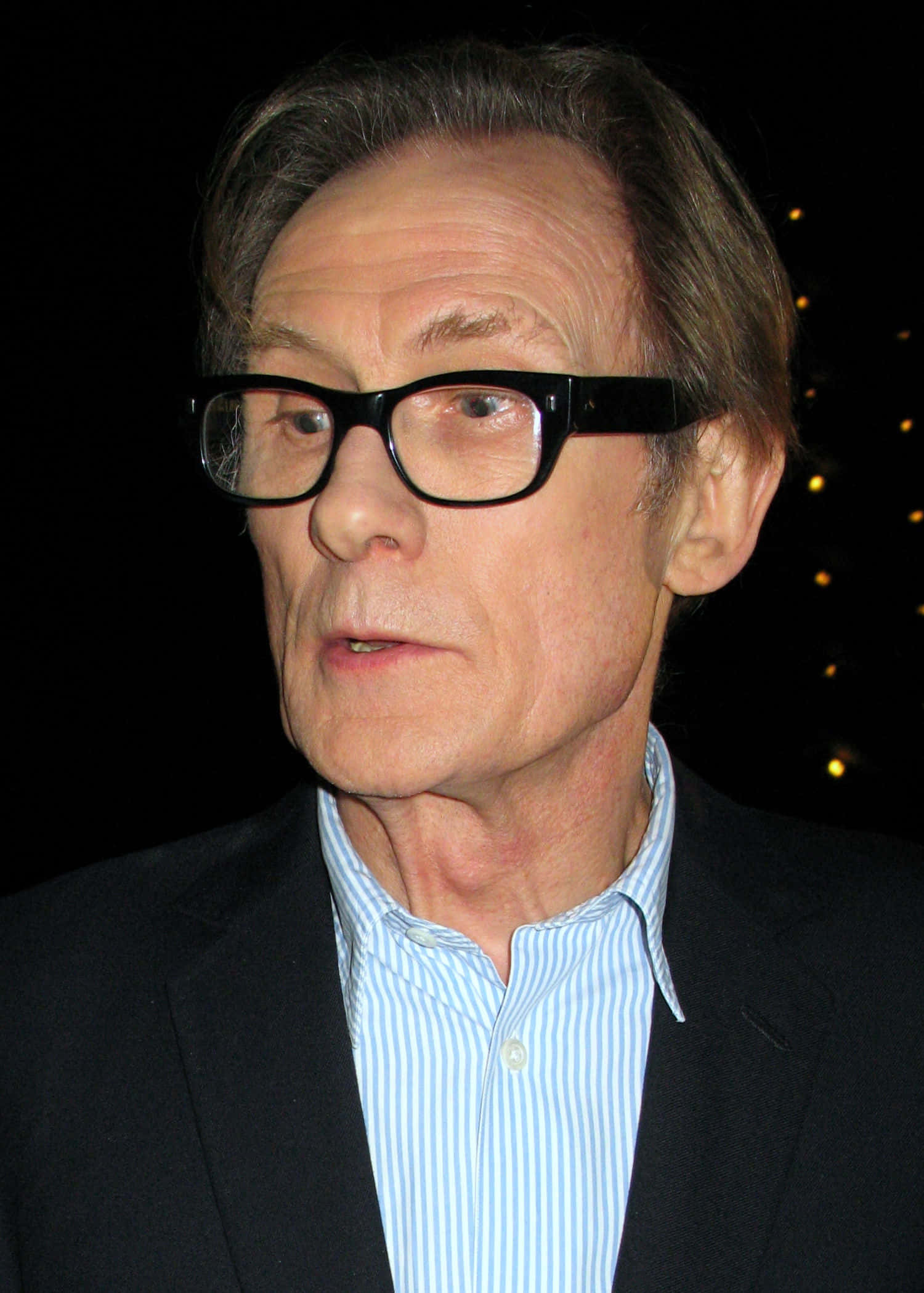 演員比爾·奈伊面部特寫 壁紙 — 一個戴眼鏡、身穿深色西裝和條紋襯衫的男士特寫肖像，背景昏暗帶有模糊燈光。