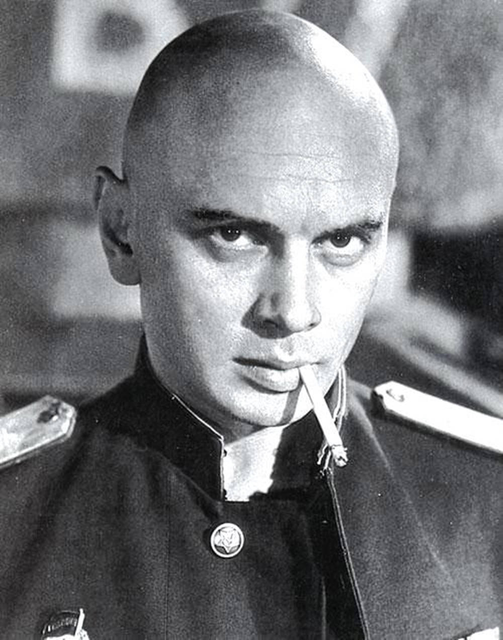 一個光頭男子穿著軍服，帶著嚴肅的表情抽煙。