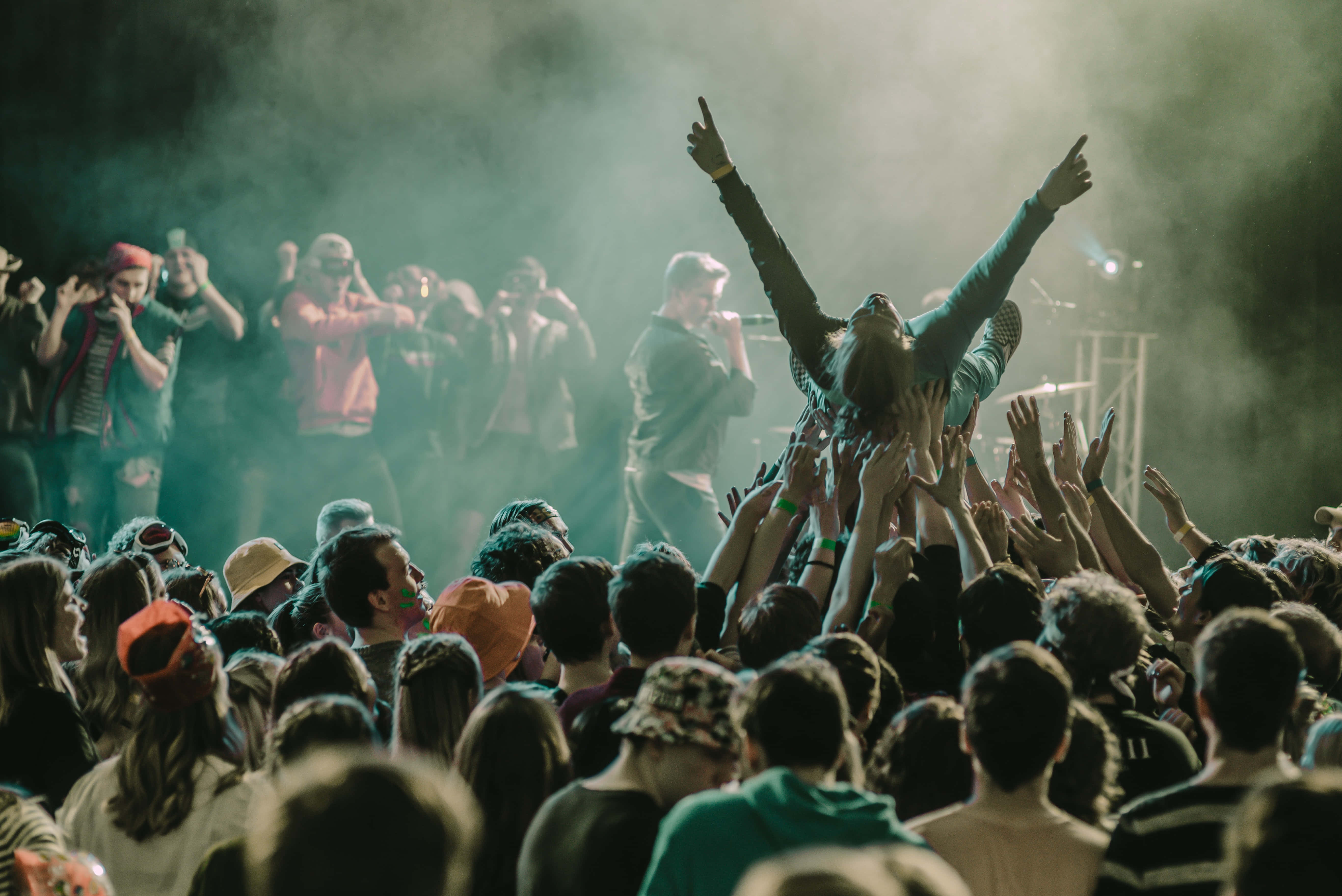 music festival 桌布 壁紙 — 演唱會上人群湧動，一名粉絲在煙霧繚繞的舞台燈光下被高舉過人群，雙臂高舉。