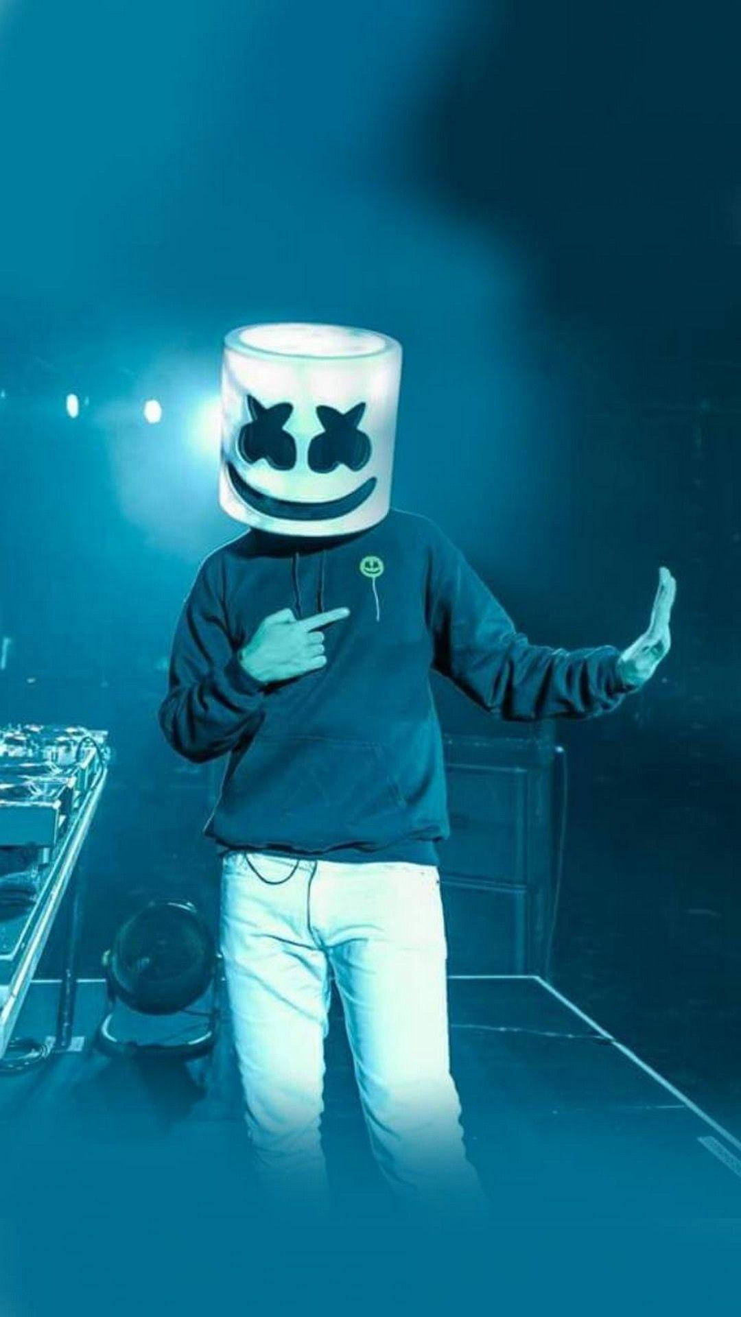 演唱會舞台Marshmello iPhone壁紙 壁紙 — 一位戴著發光白色棉花糖頭盔的DJ在藍色燈光、煙霧和設備的舞台上自信地指著。