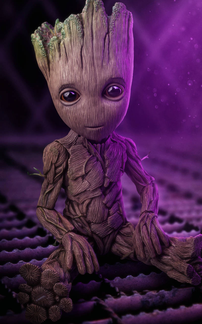 漫威小樹人寶寶 壁紙 — 一個細小嘅樹狀生物，有大眼睛，平靜咁坐喺紫色紋理表面，喺發光紫色燈光下。