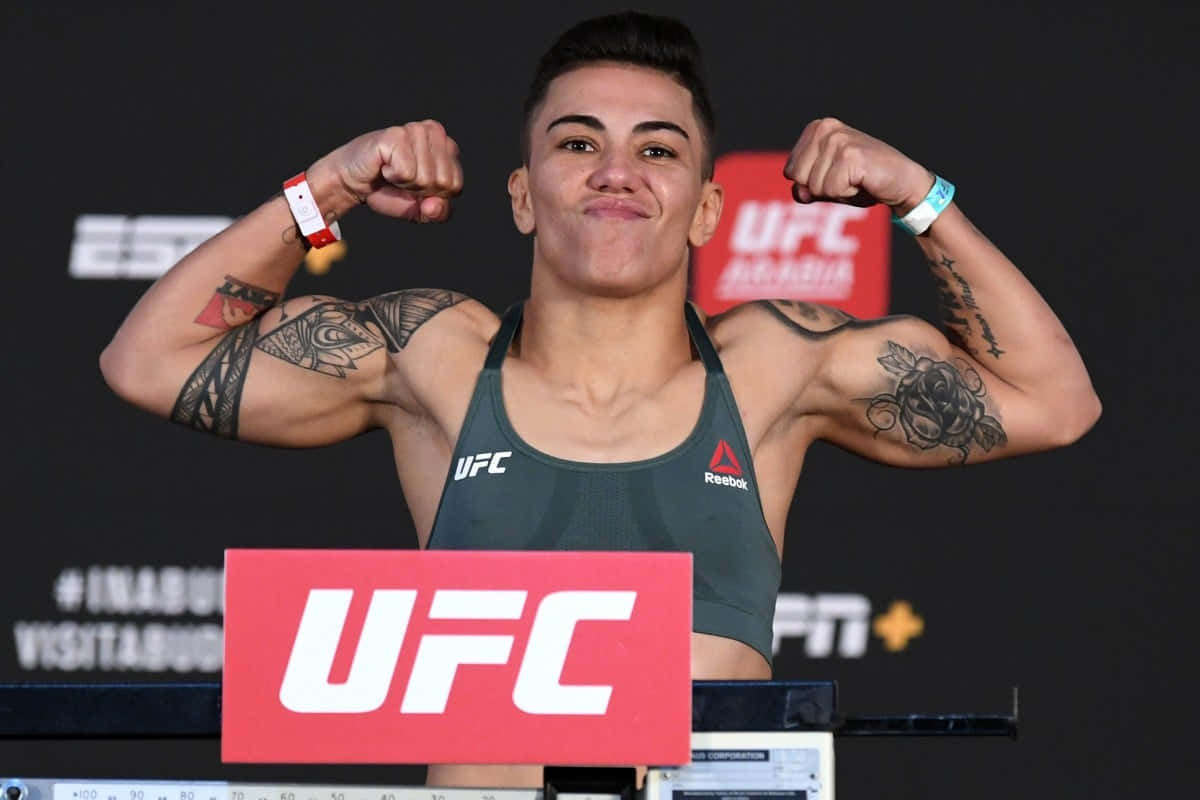 女性UFC拳擊手在稱重活動中彎曲手臂，帶有紋身和品牌標誌。