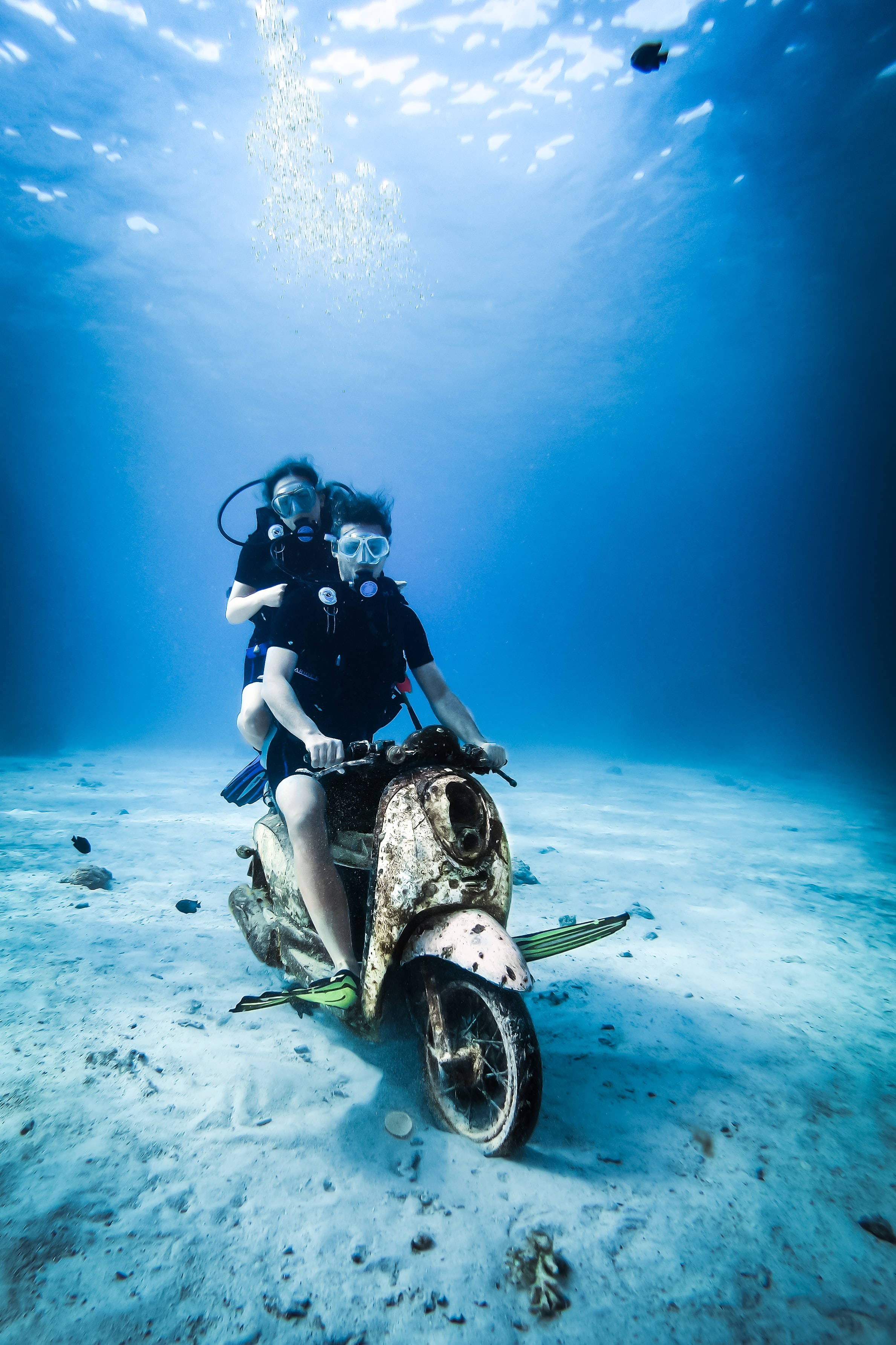 潛水與水下摩托車合影 壁紙 — 兩名水肺潛水員在明亮的藍色海水下，騎著一輛生鏽的摩托車潛伏在沙質海床上。