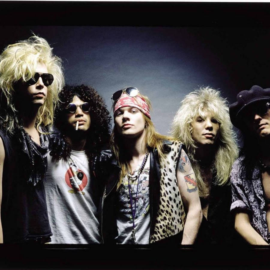 潮汐音樂會槍與玫瑰演出 壁紙 — Guns N' Roses 樂隊成員以狂野的頭髮、太陽眼鏡和皮背心在黑暗的錄音室背景下擺姿勢。