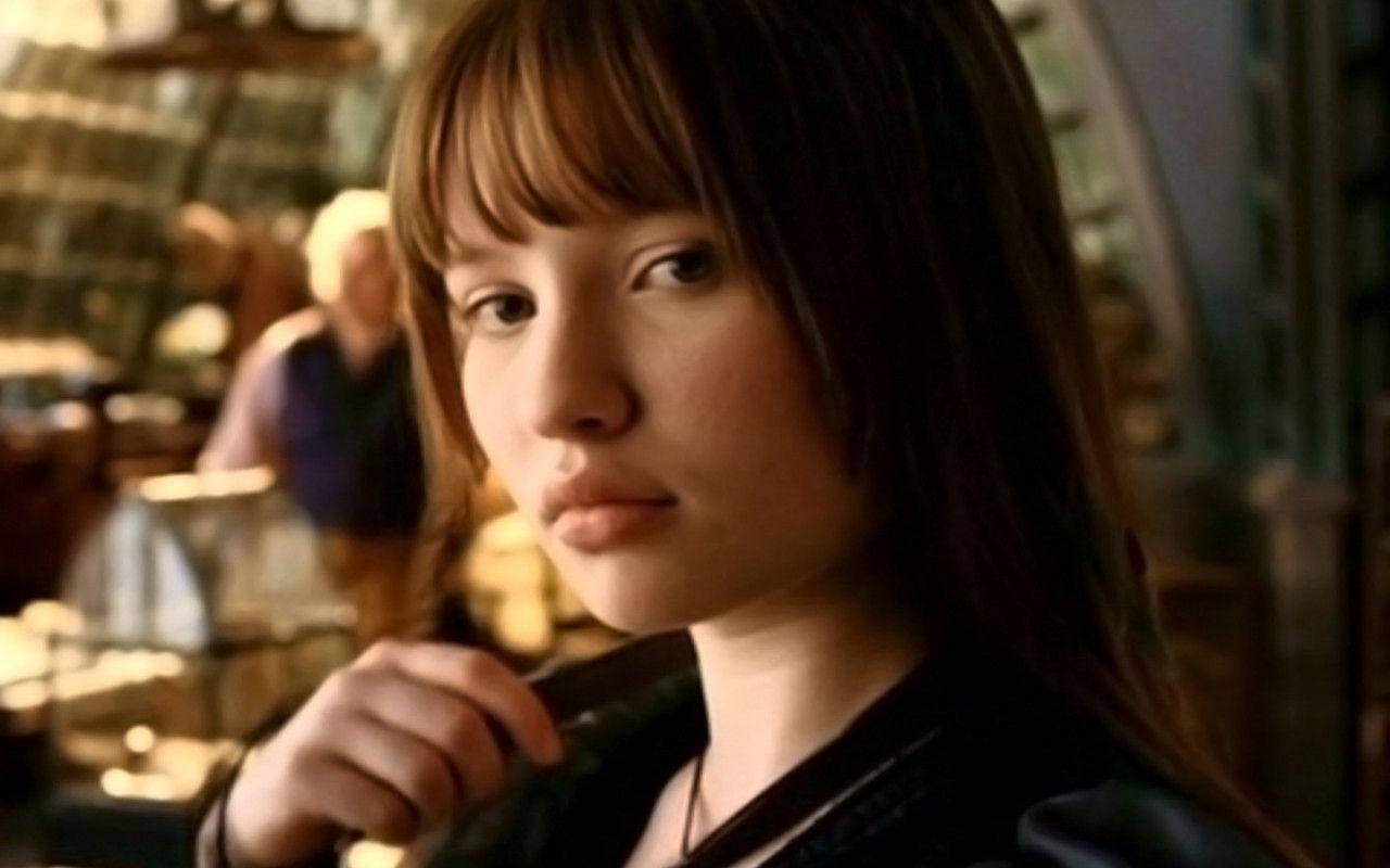 澳洲女演員艾米莉·布朗寧飾演《Lemony Snicket's A Series of Unfortunate Events》中的Violet Baudelaire（2004） 壁紙 — 一位棕色頭髮和瀏海的年輕女性在溫暖照明的室內環境中直視相機，背景人物模糊。