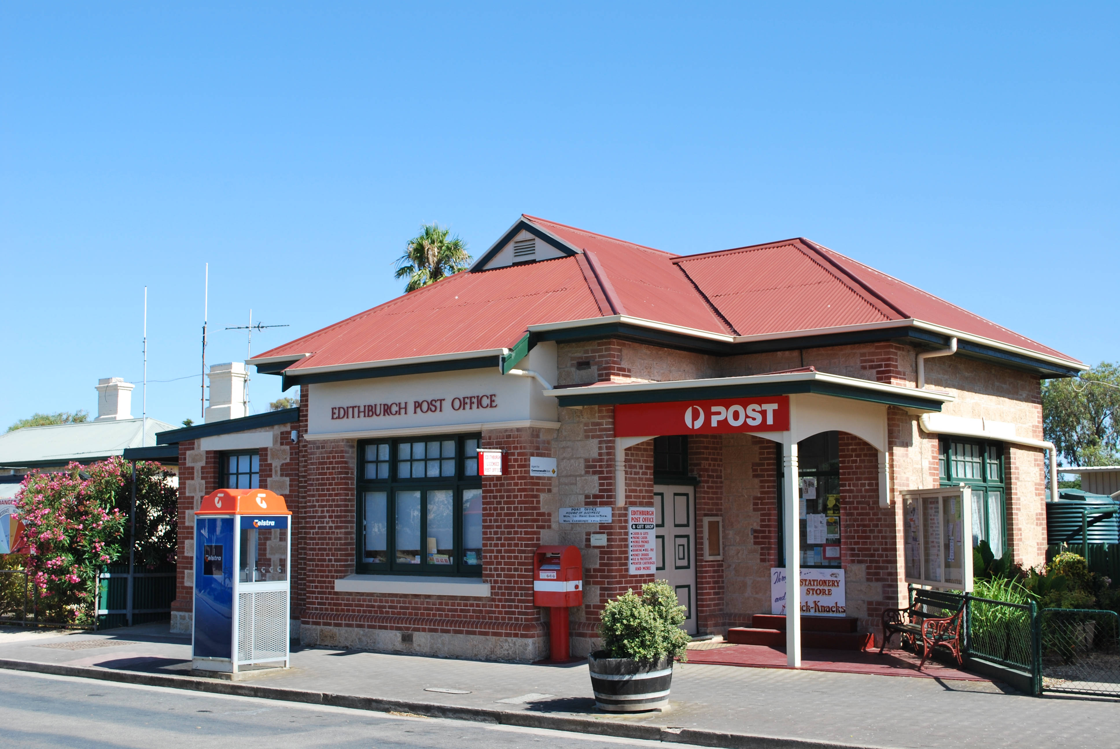 澳洲愛迪斯堡郵政局 壁紙 — 一座明亮的紅瓦磚造郵局，帶有「EDITHBURCH POST OFFICE」標誌，矗立在晴朗的藍天下，附近有盛開的粉紅色花朵。