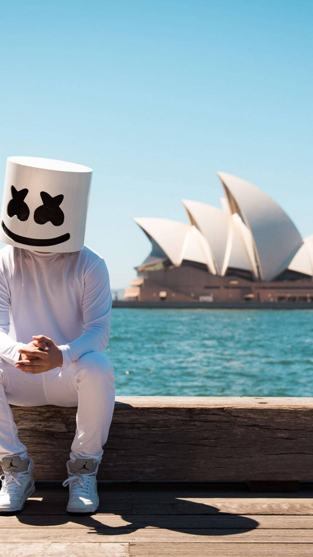 澳洲歌劇院Marshmello iPhone壁紙 壁紙 — 一個人戴著白色棉花糖面具和穿著白色棉花糖服裝，坐在木製碼頭上，背景是悉尼歌劇院和藍色海水，天空晴朗。