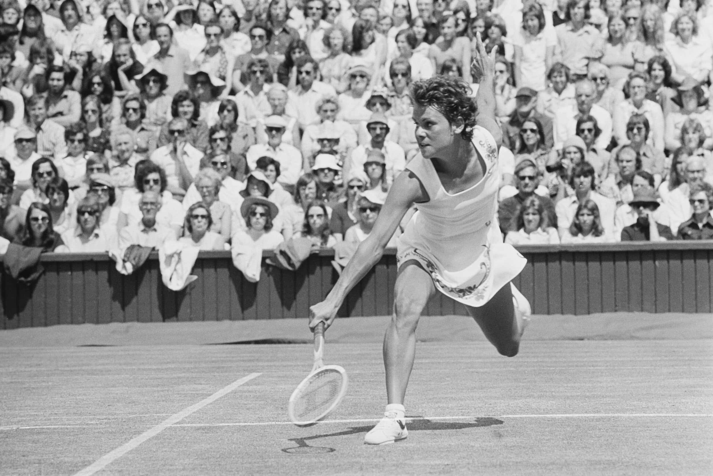 澳洲網壇傳奇 Evonne Goolagong Cawley 動態賽場黑白快照 壁紙 — 一位女網球手在陽光明媚的球場上揮拍中途向前猛衝，人群在黑白畫面中專注觀看。