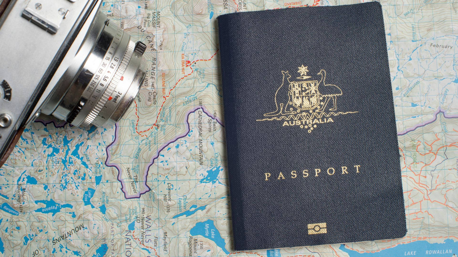 澳洲護照與相機置於世界地圖上 壁紙 — 一本海軍藍色的澳洲護照放在地形圖上，旁邊有一部準備好旅行的復古銀色相機。