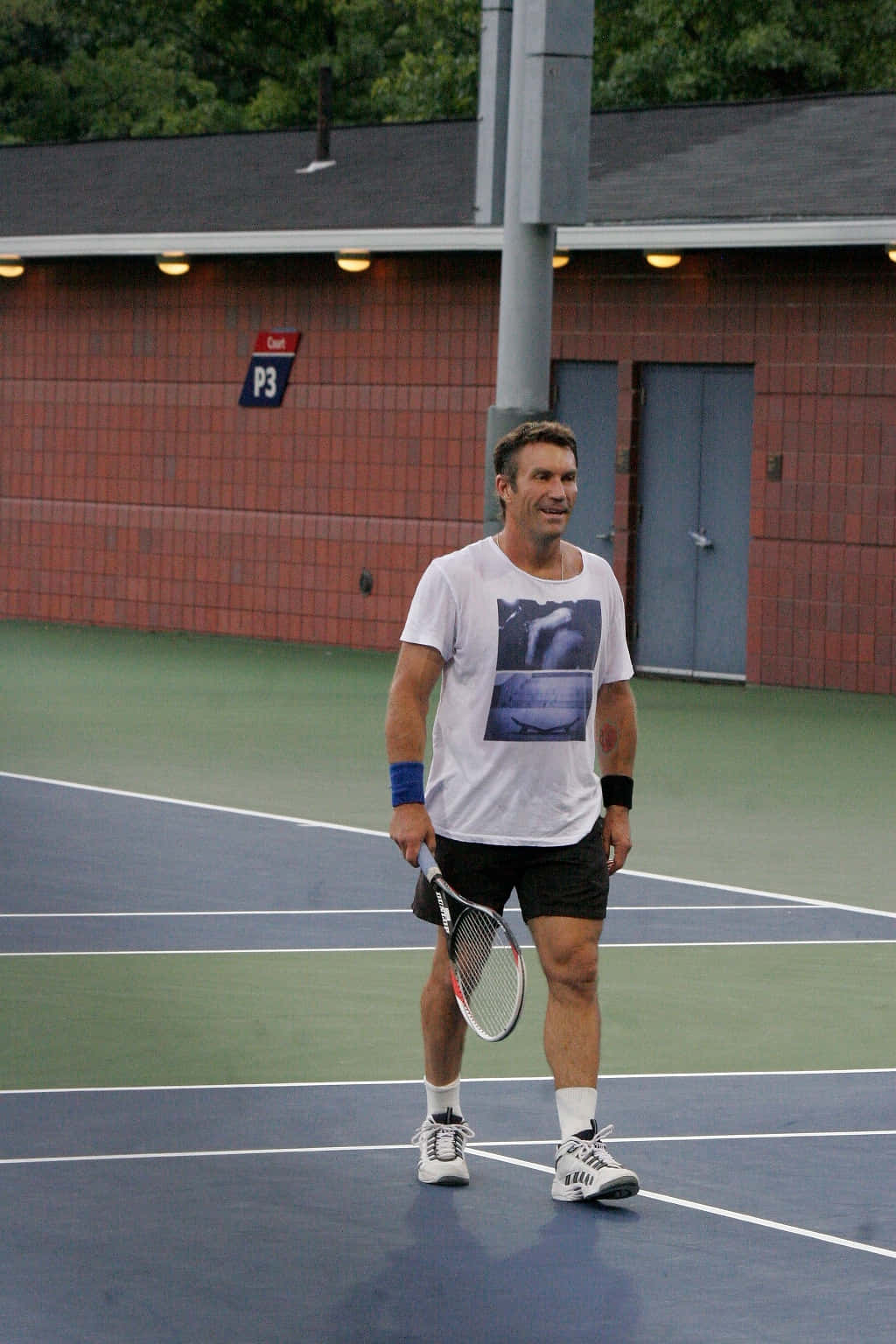 澳洲運動員 Pat Cash 壁紙 — 一個微笑嘅男人著住運動衫企喺藍色網球場上，揸住球拍，背景有磚樓同樹。
