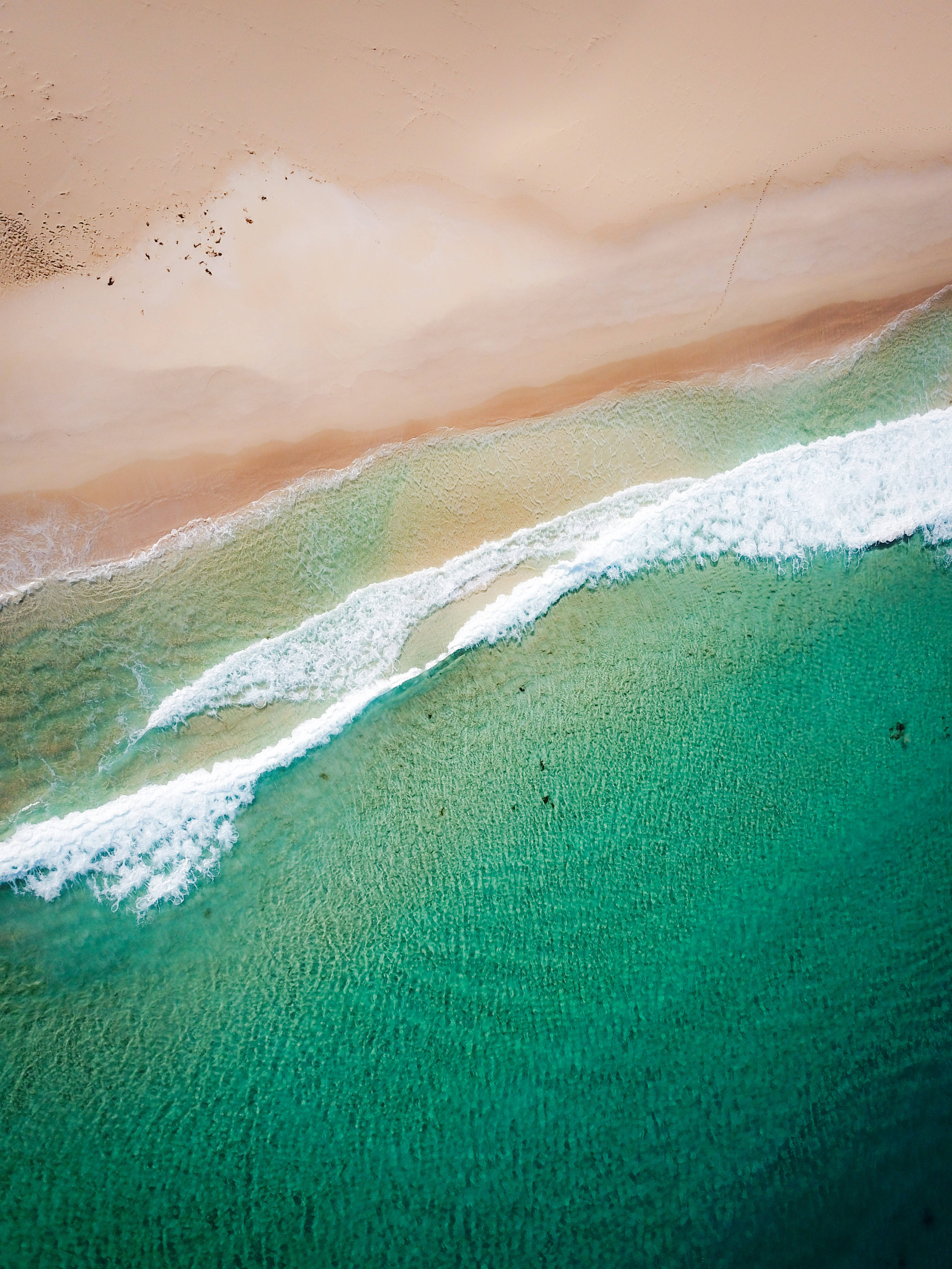 澳洲邦迪海灘 iPhone 背景 壁紙 — 從高空俯瞰，青綠色的海浪拍打著柔軟的蜜桃色沙灘，呈現出清晰的水紋圖案。