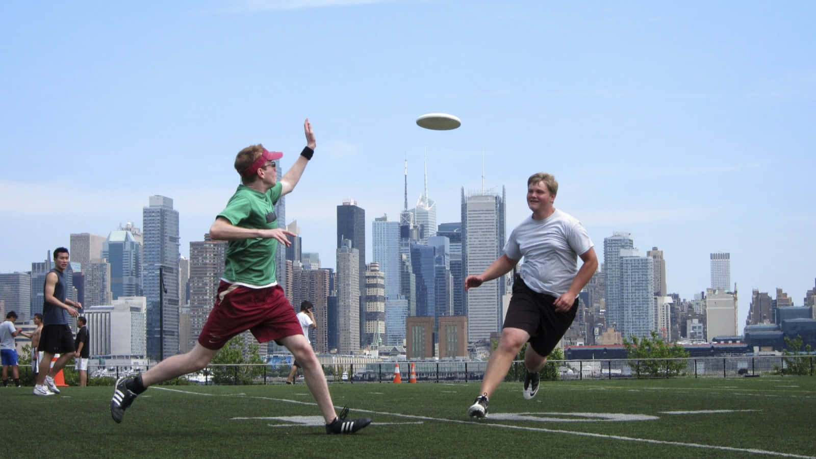 激烈 Ultimate Frisbee 比賽實況 壁紙 — 兩個男人跳起伸手去接一個飛行的飛盤，在一個有草皮的屋頂場地上，背後是高聳的城市天際線。