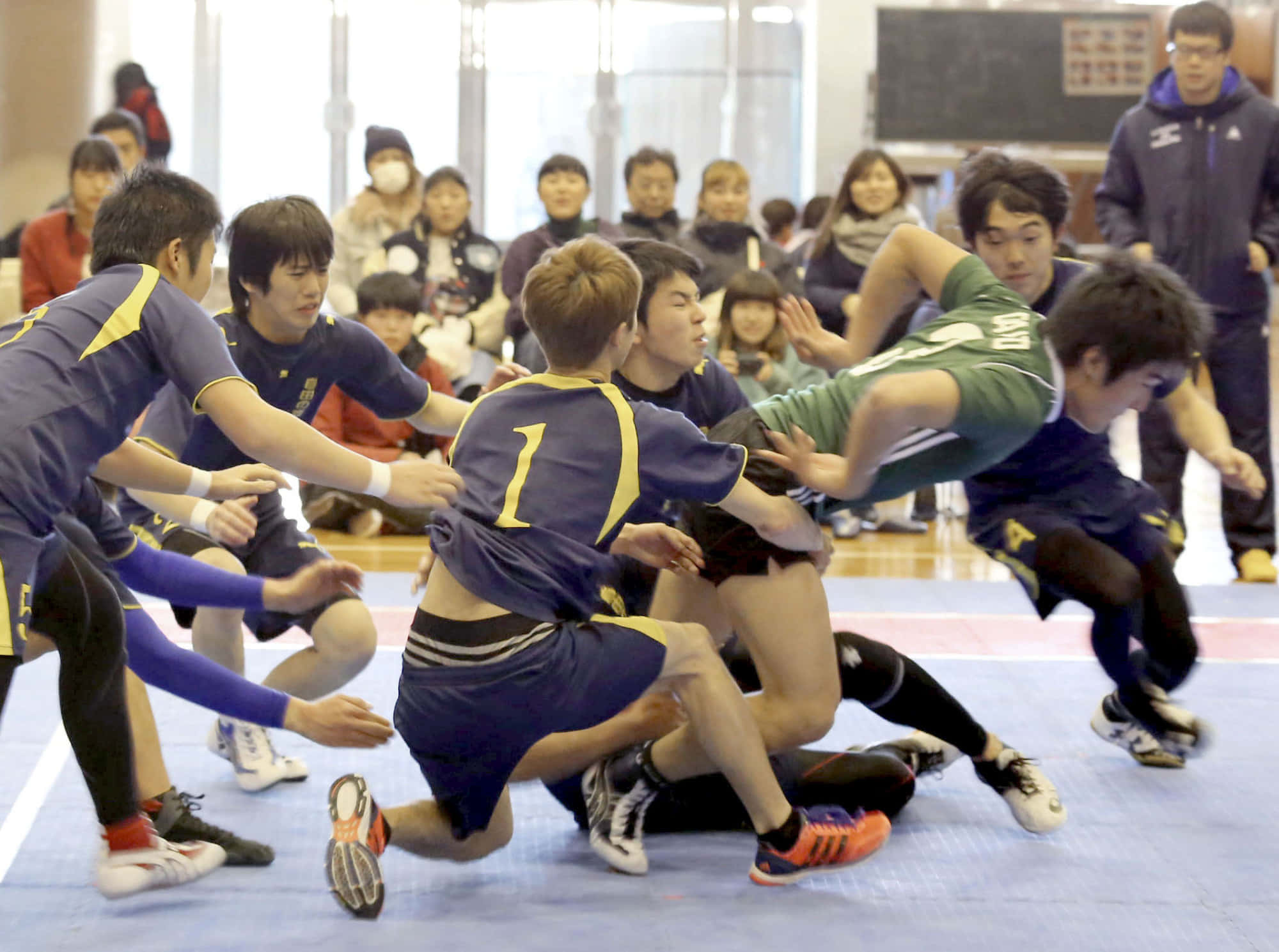 Kabaddi 桌布 壁紙 — 激烈的室內欖球比賽，球員身穿藍色和綠色球衣，在藍色墊子上激烈纏鬥，觀眾在場邊觀看。