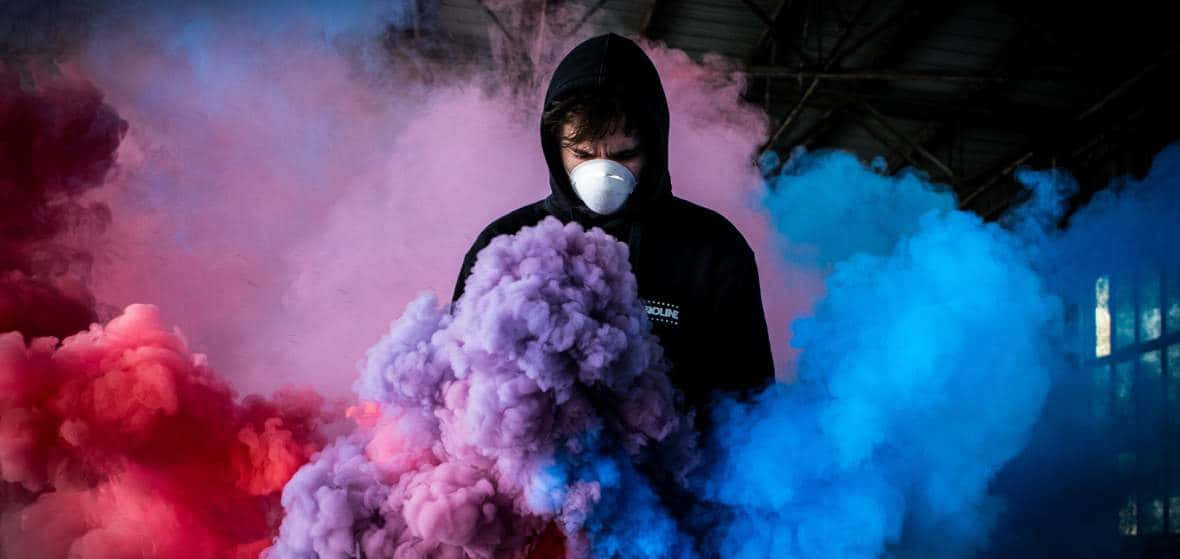 在黑暗的工業環境中，穿著黑色連帽衫和白色口罩的人置身於彩色煙霧中。
