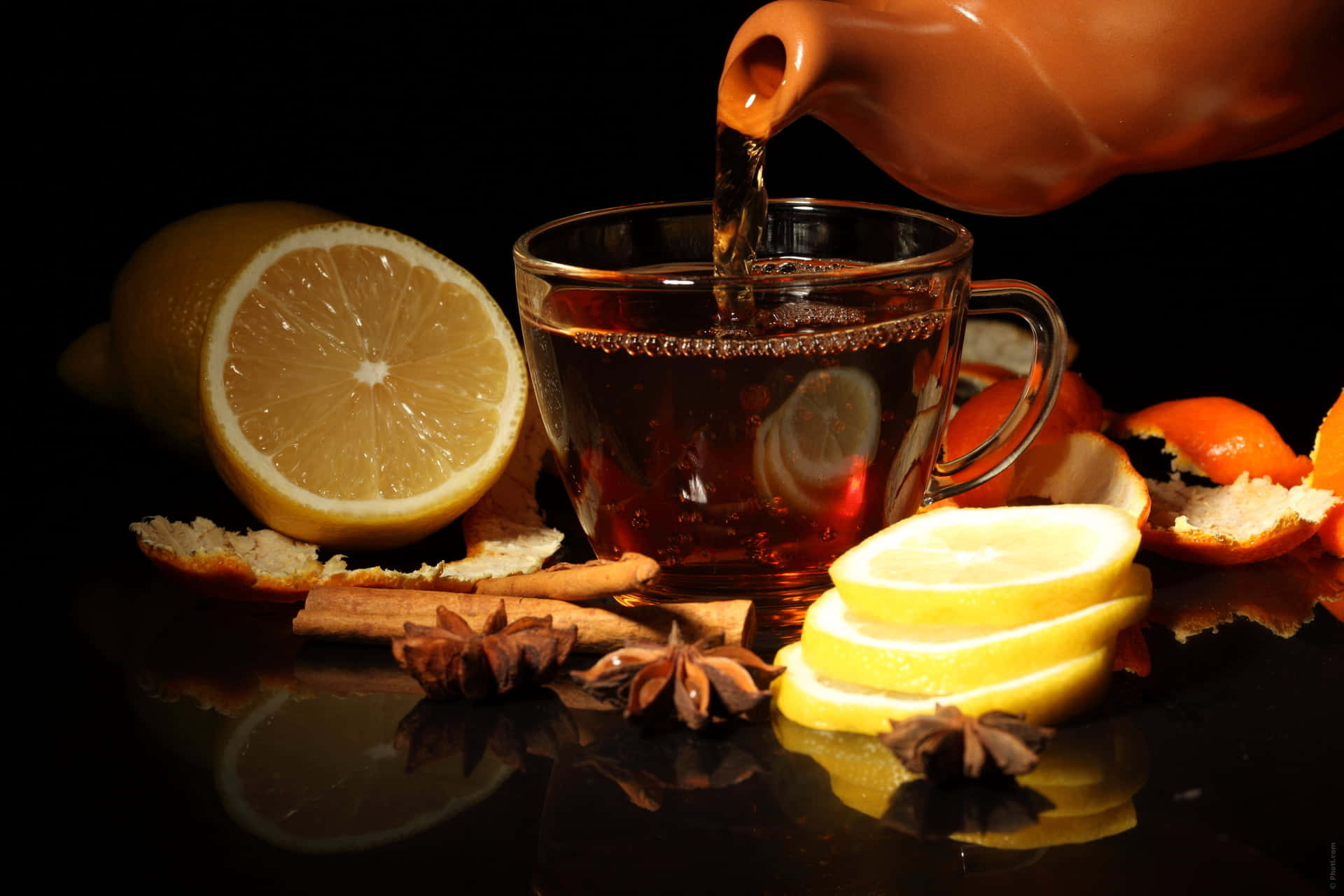 Mulled Wine 桌布 壁紙 — 濃郁琥珀色茶從陶瓷壺倒入玻璃杯中，周圍有檸檬片、八角及完整柑橘類水果，置於深色反光表面上。