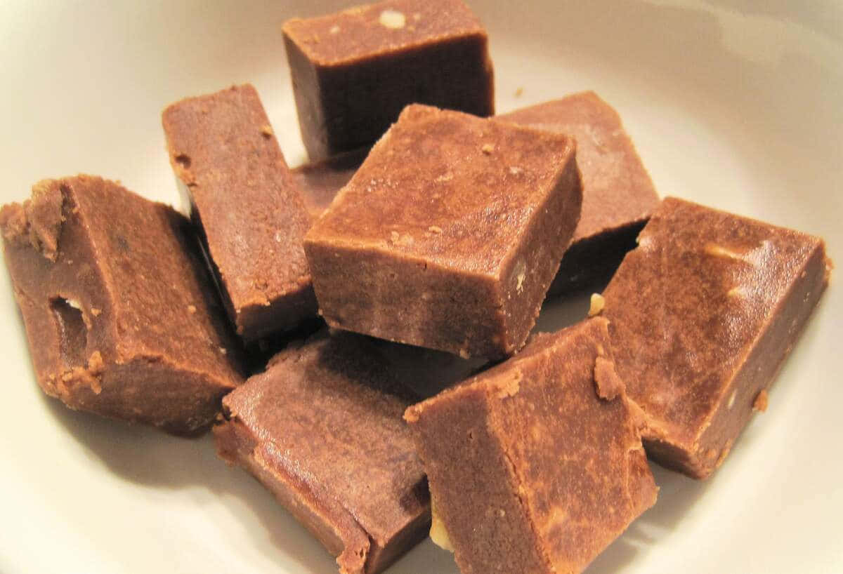 Penuche Fudge 桌布 壁紙 — 濃郁的棕色軟糖方塊誘人地堆疊在白色盤子上，帶有可見的堅果碎塊和光澤質感。