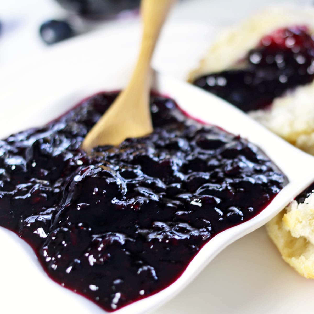 Blueberry Jam 桌布 壁紙 — 濃郁的深色藍莓果醬在白色碟子中閃閃發光，配以木勺，旁邊是金黃色的司康餅，上面有一勺果醬。